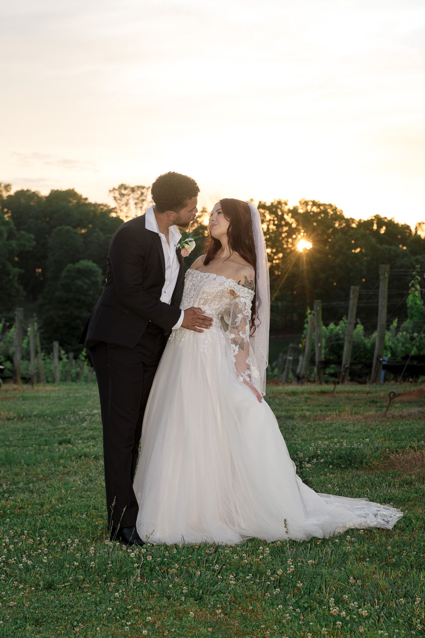 Bride and groom share a romantic sunset kiss in a vineyard, bride wearing an off-shoulder lace wedding gown with veil.