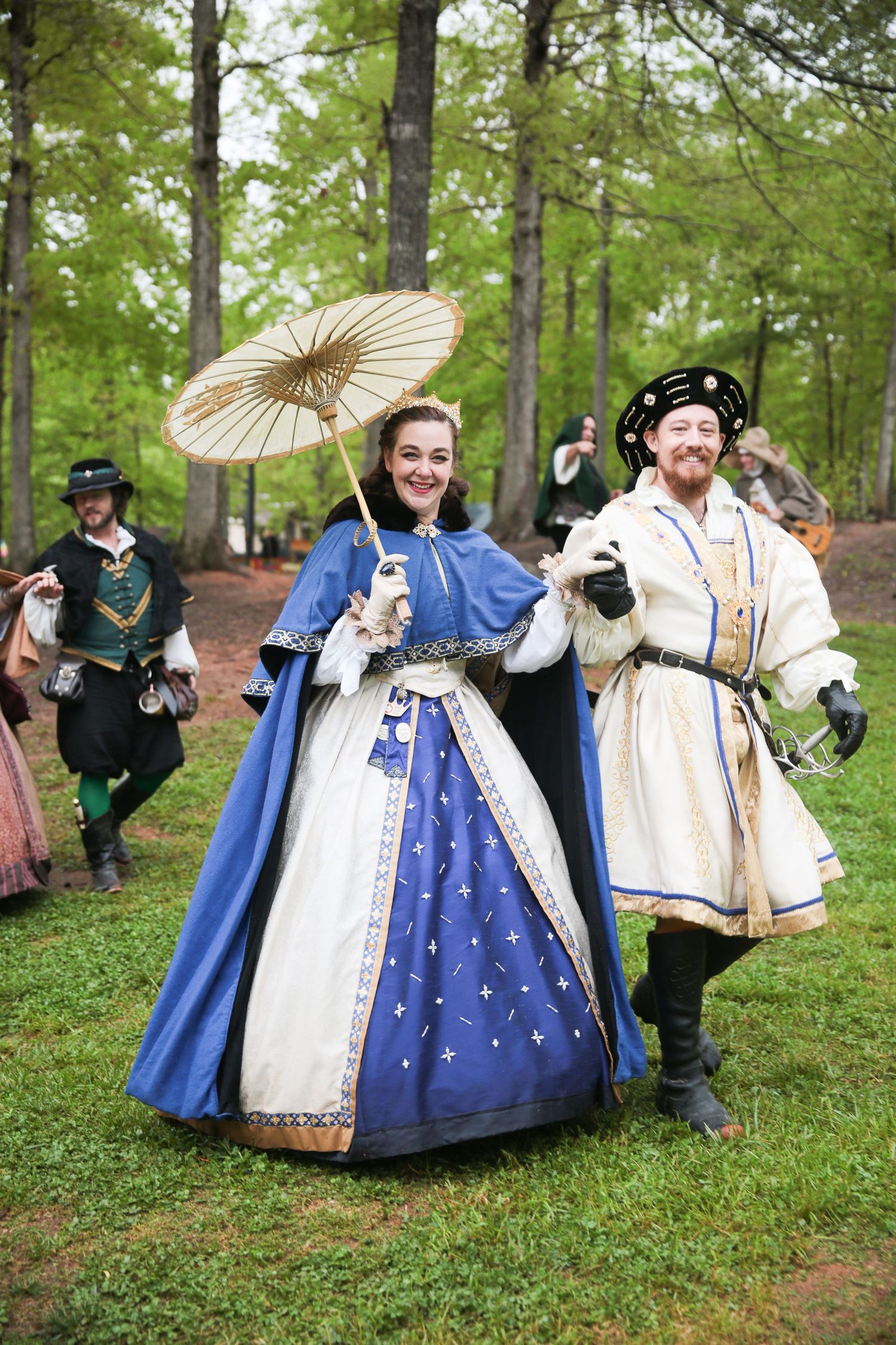Renaissance fair performers in elaborate period costumes posing with parasols on grassy grounds.