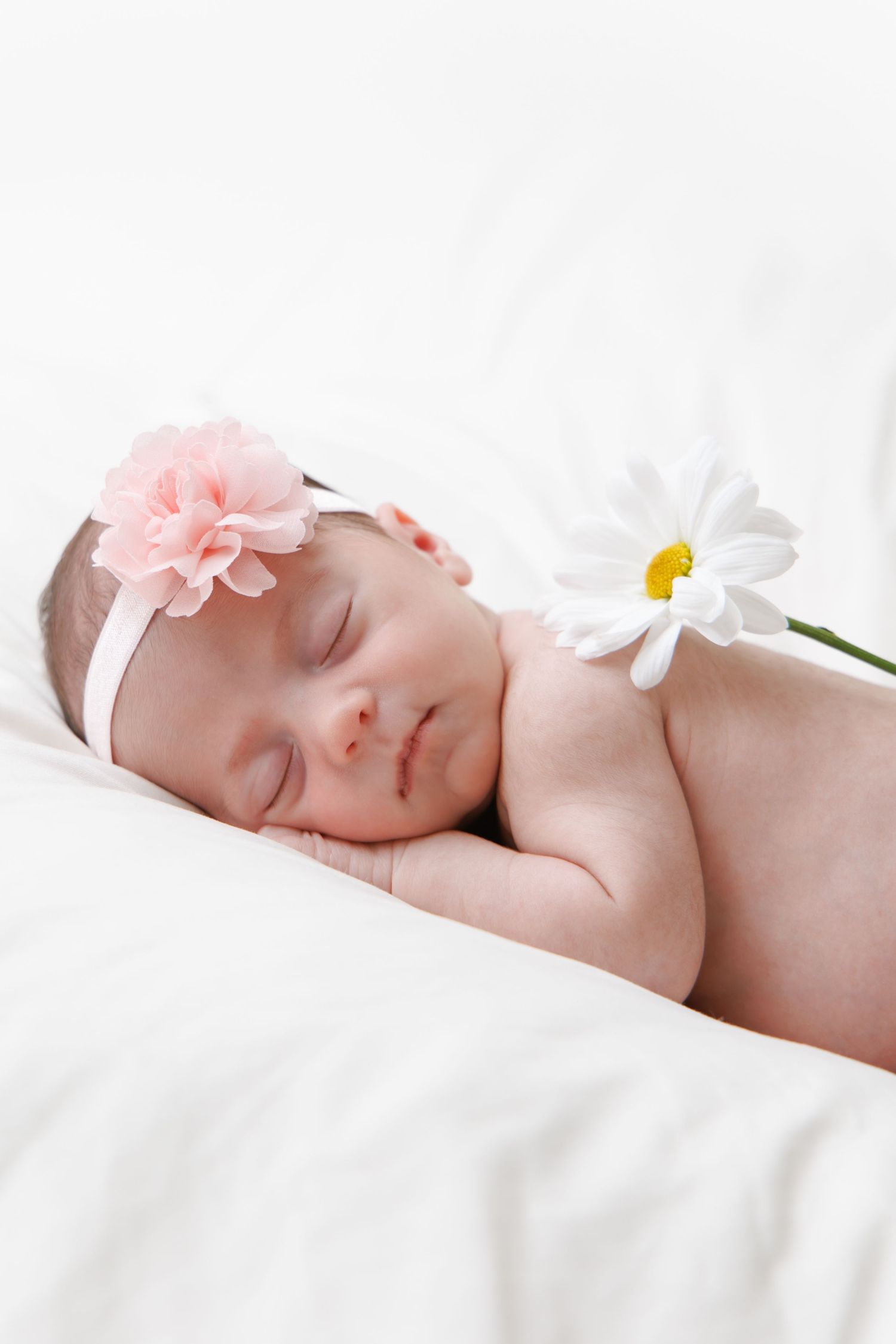 Sleeping baby in white bedding with pink flower accessory and white daisy flower decoration.