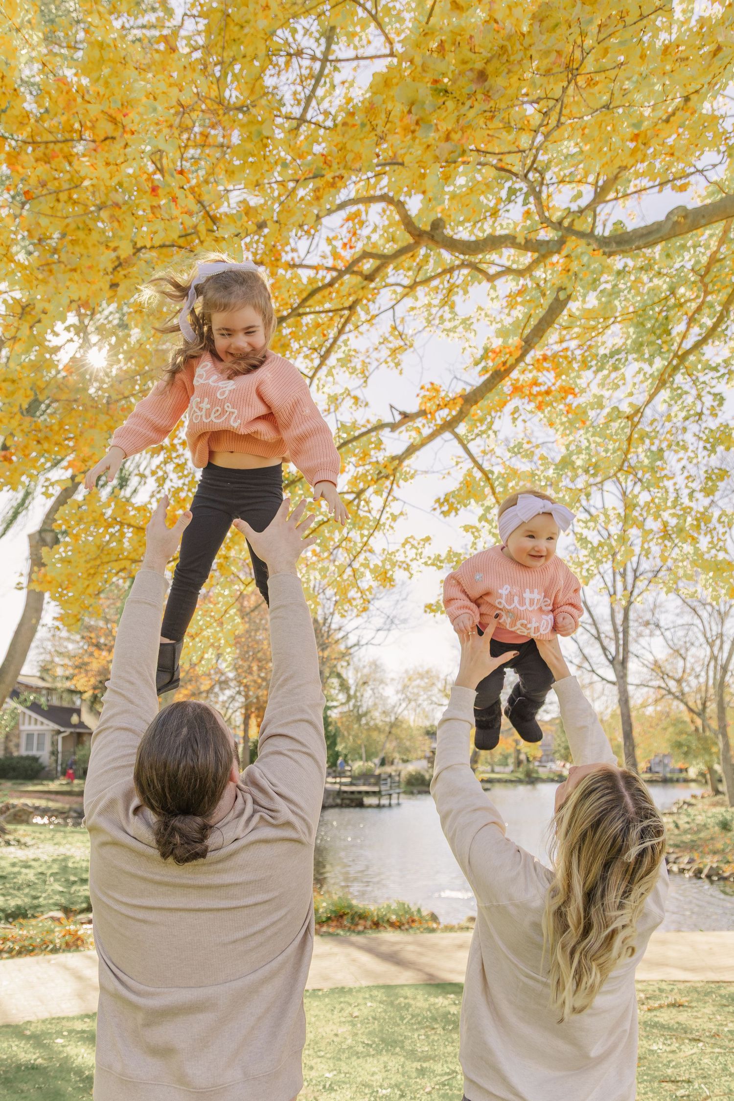 Two adults lift children wearing matching coral shirts under vibrant yellow autumn trees in a park setting.
