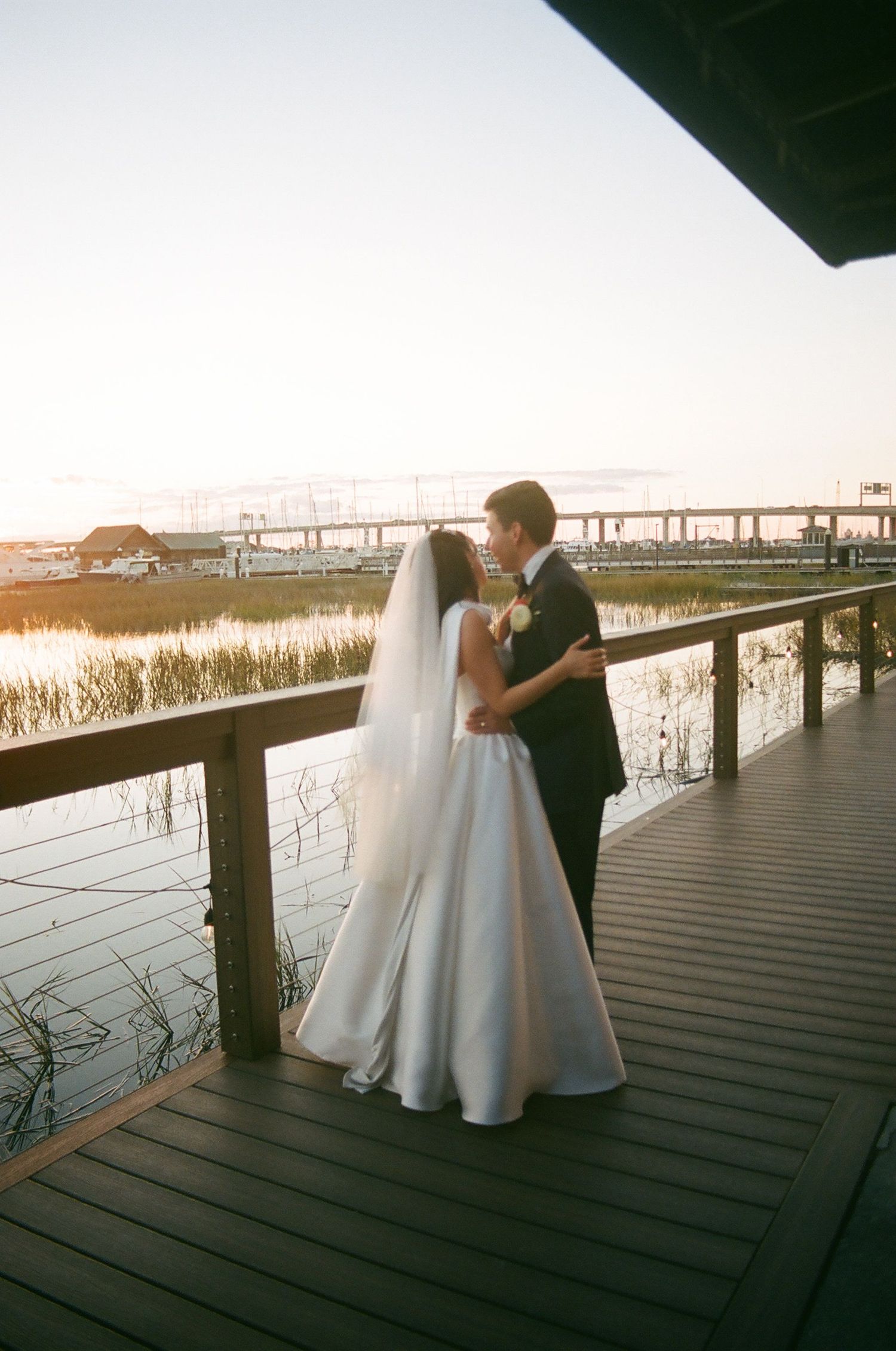 A couple shares a romantic moment on a wooden deck overlooking a marsh at sunset.