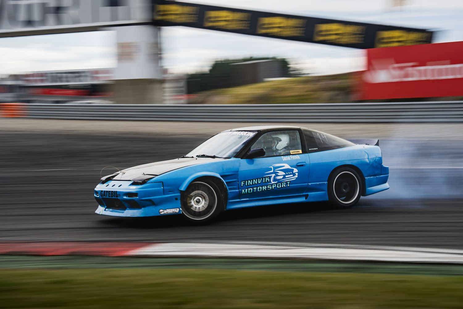 A blue drift car performs a high-speed turn on a race track with smoke trailing from its tires.