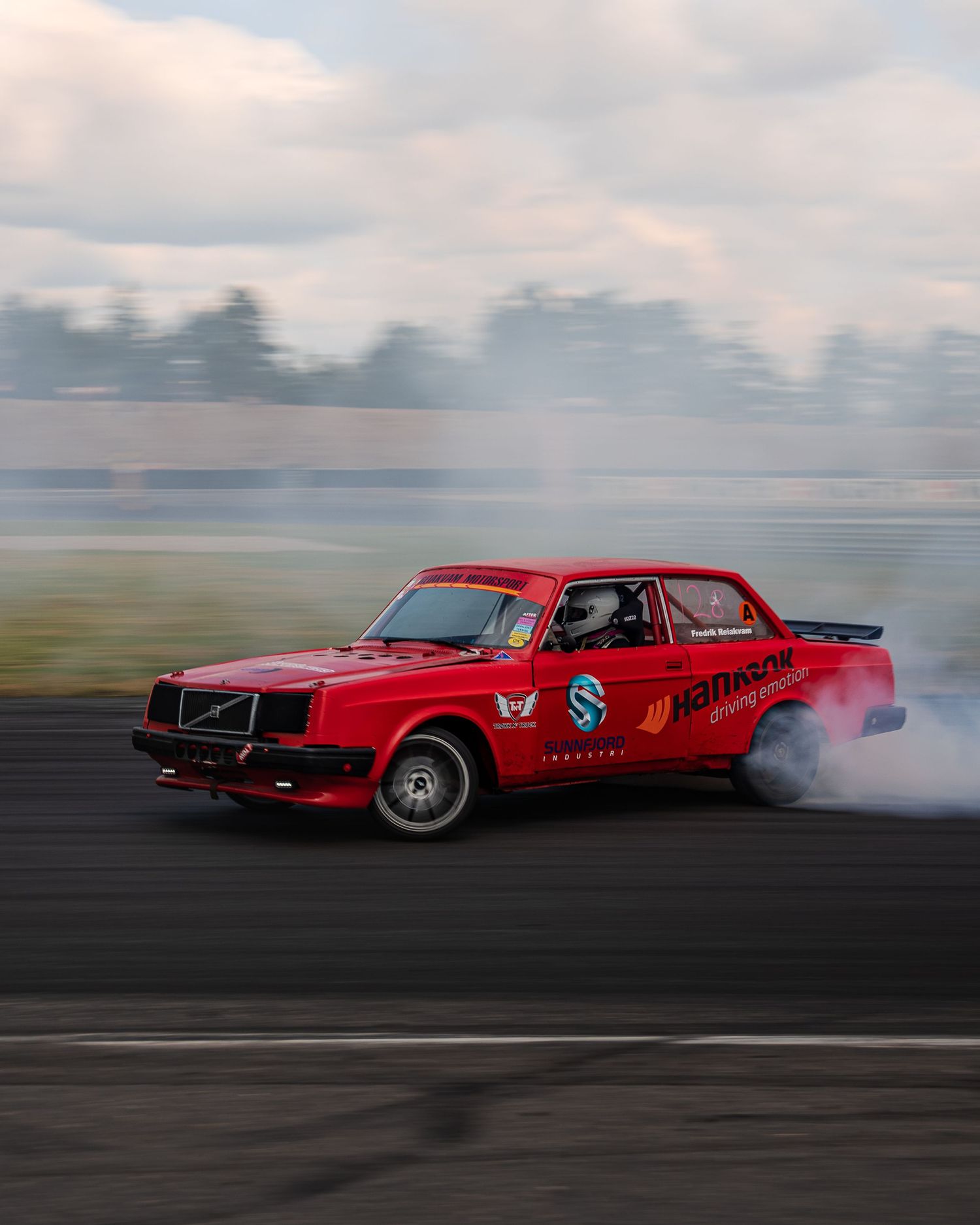 A red vintage drag racing car performing a burnout on a track, creating dramatic smoke trails behind it.