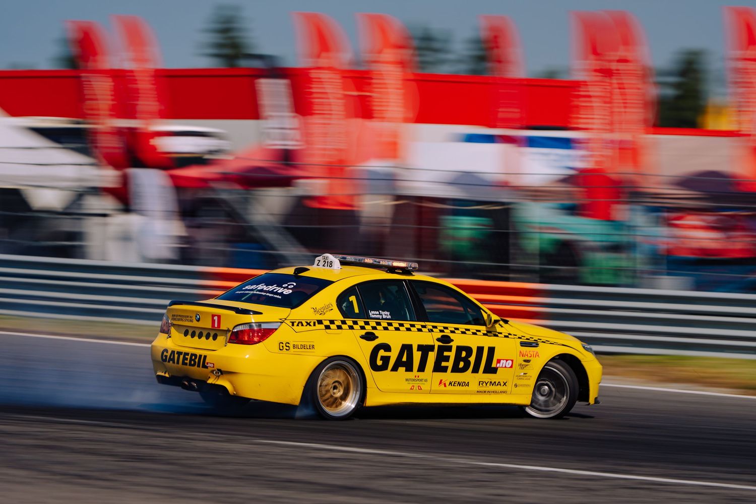 Yellow taxi racing cars speed past on a racetrack with red barriers and spectator stands visible in the background.