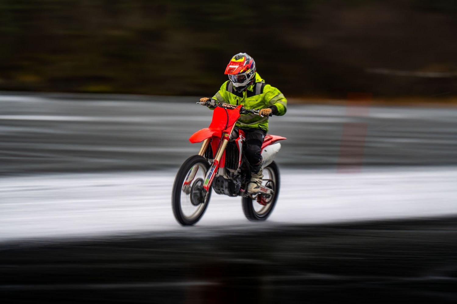 Dirt bike rider in bright safety gear speeds through a motion-blurred forest track during an off-road race.