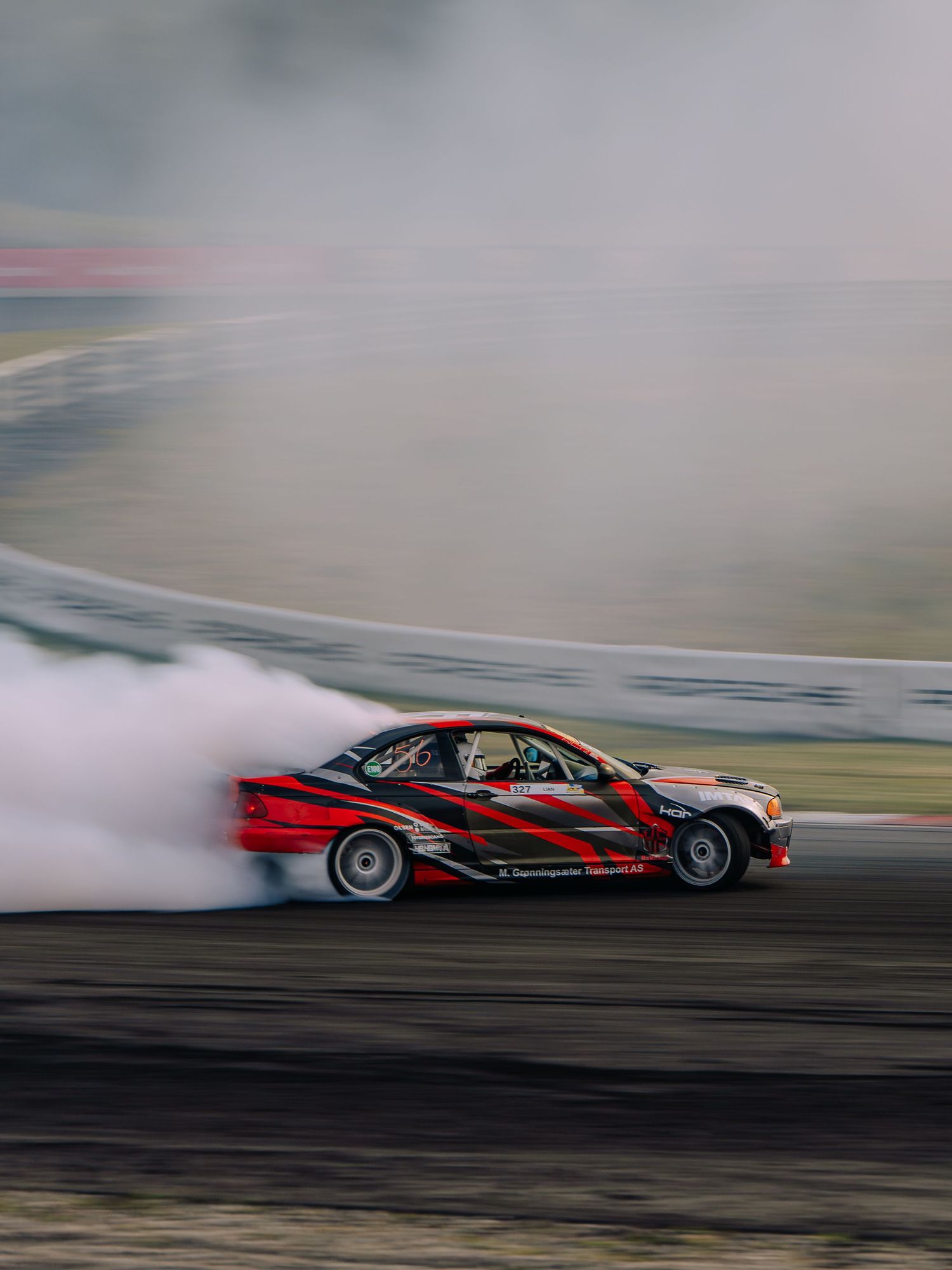 Red BMW drifting on a racetrack creating smoke while performing a controlled slide during motorsport competition.