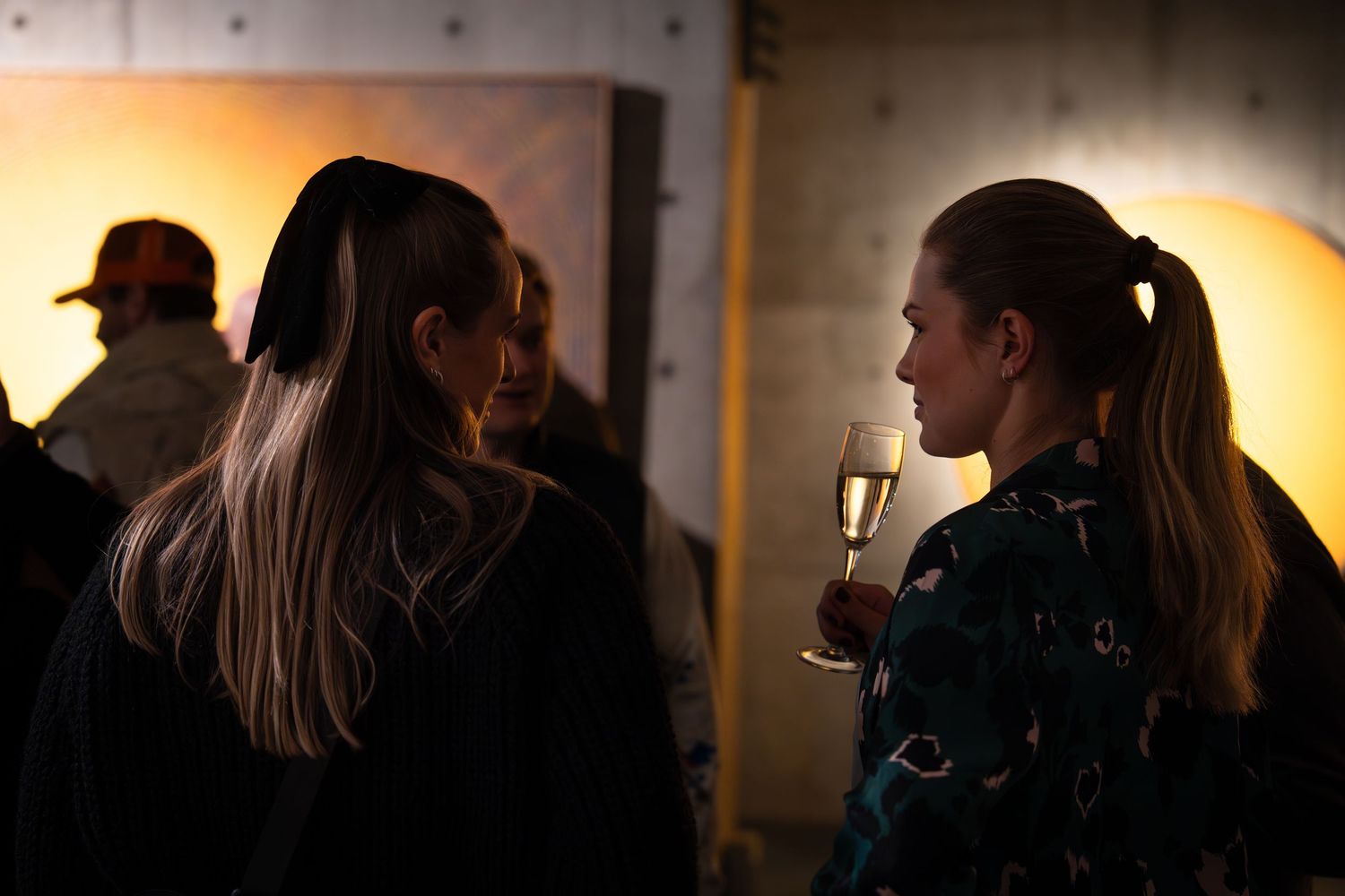 Art gallery attendees view illuminated artwork in a dark exhibition space.