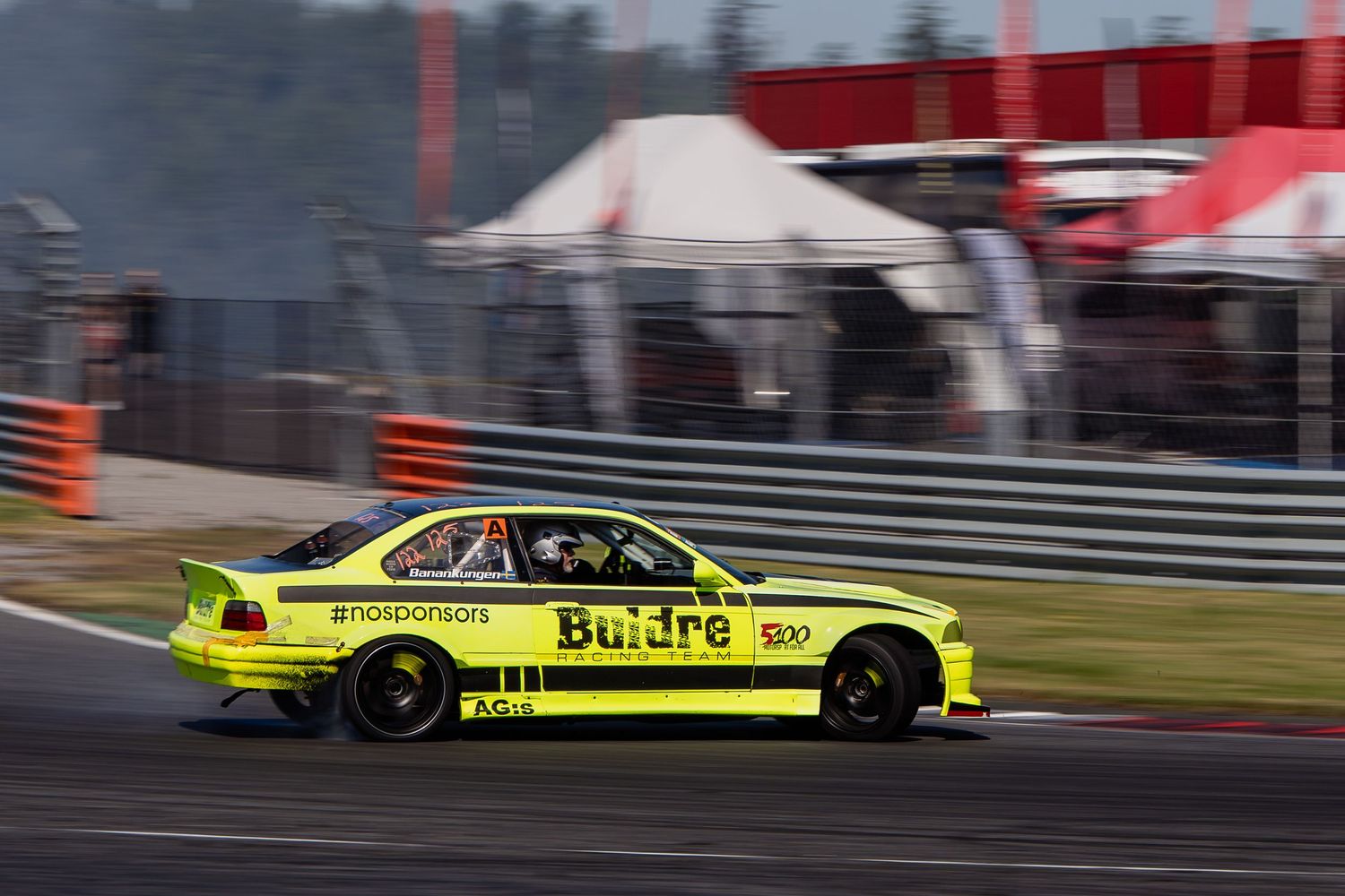 A yellow race car drifts sideways on a race track with smoke trailing from its tires during a motorsport event.