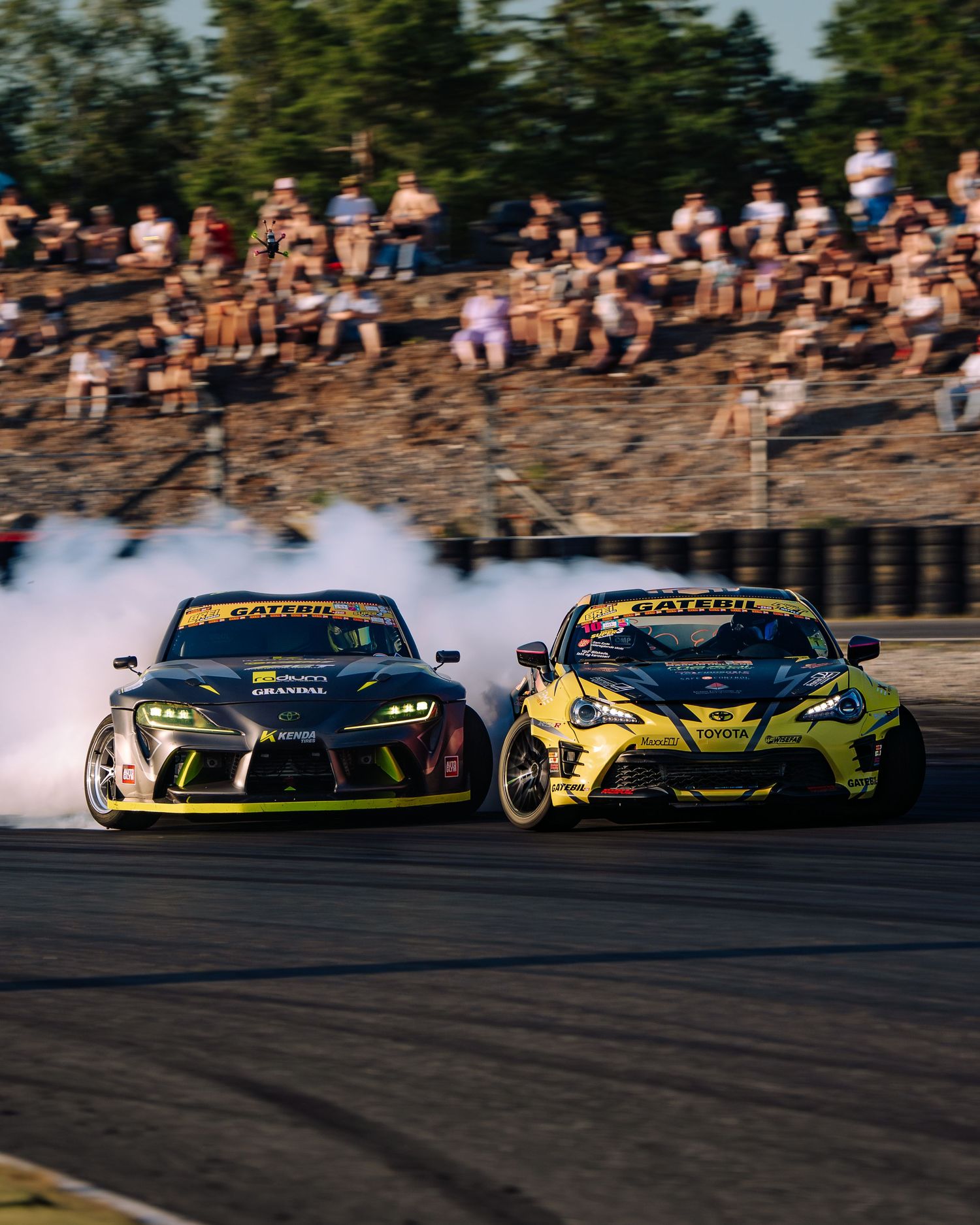 Two drift cars, one black and one yellow, racing side by side with smoke on a track as spectators watch from the hill.