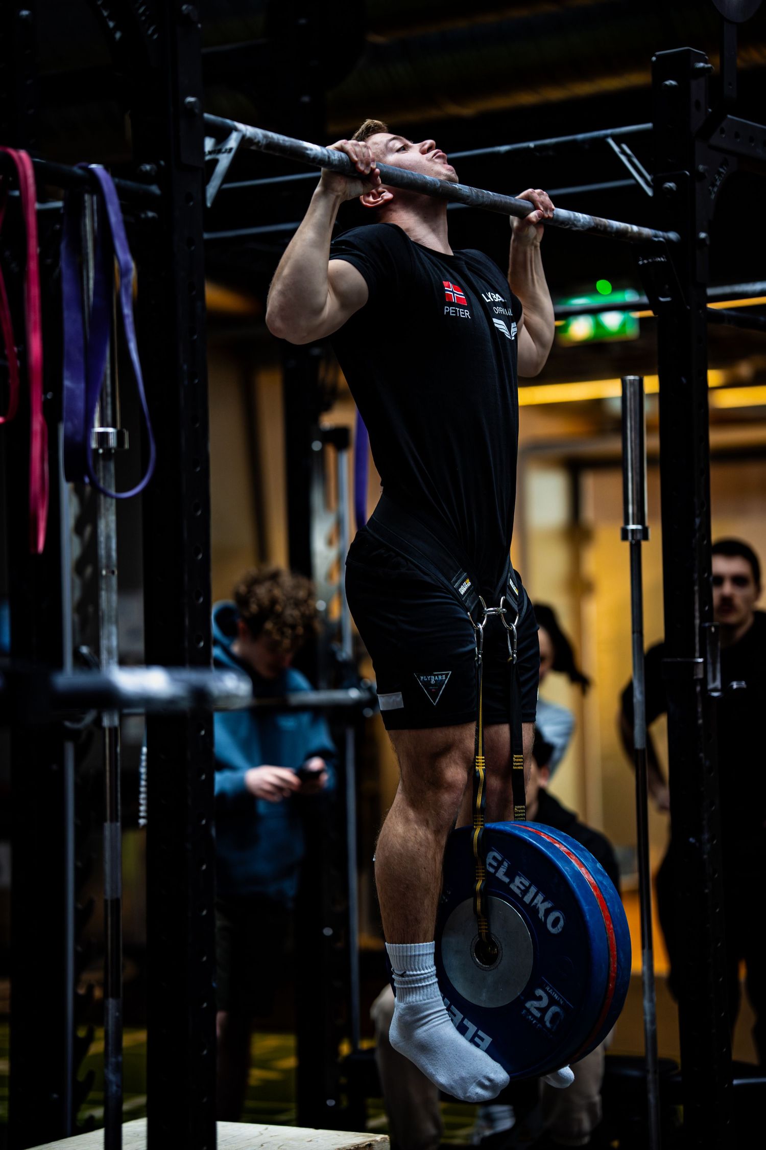 Athlete performs a pull-up on a gym bar with a weight plate attached to their belt in a dark gym setting.