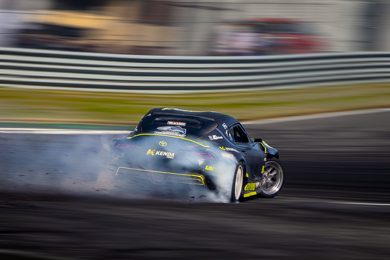 A drift car slides sideways on a race track creating a cloud of tire smoke during a competitive drift event.