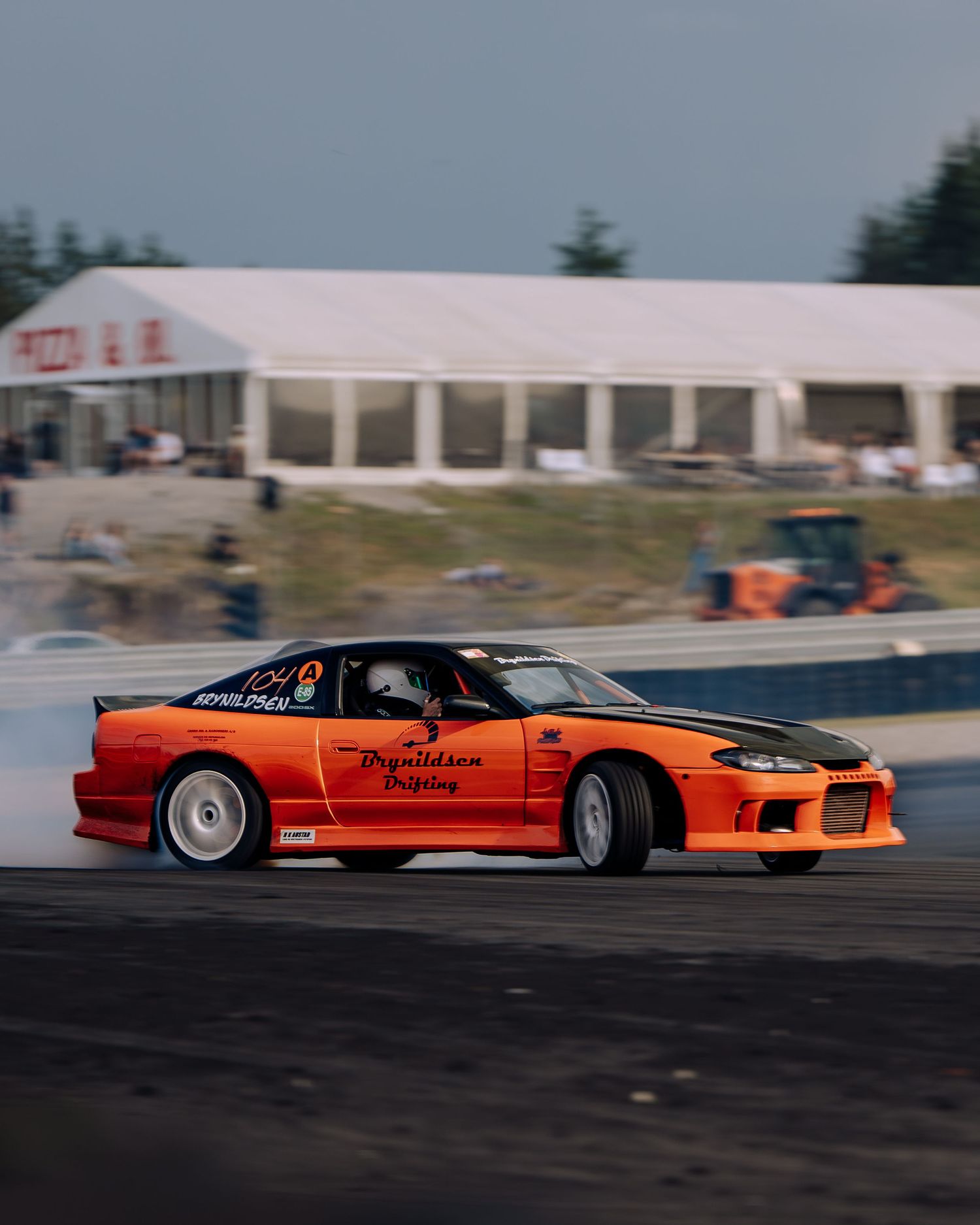 An orange drift car performs a high-speed slide on a racetrack, creating dramatic smoke from the tires.