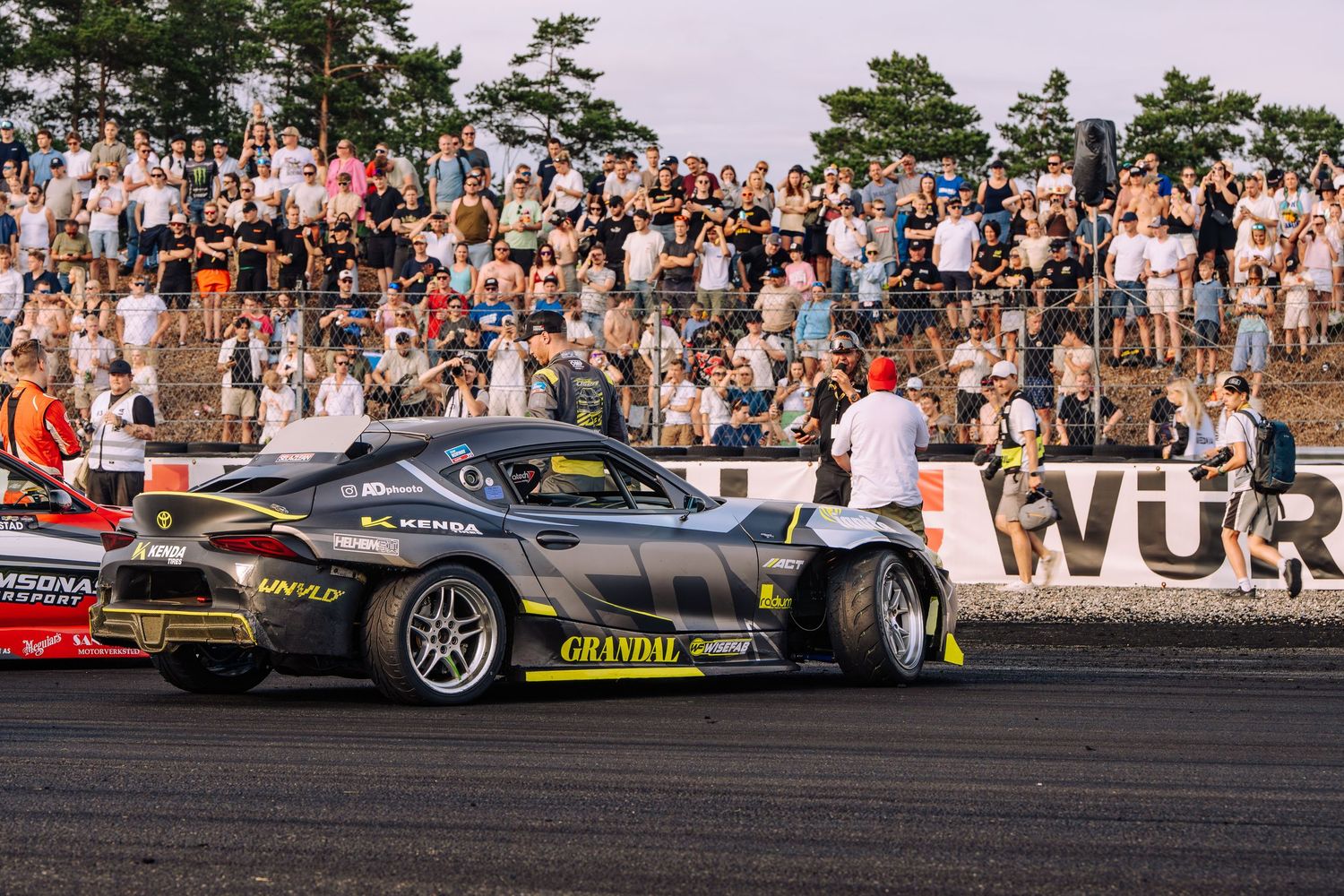 A drift car performs at a motorsport event with a large crowd watching from bleachers in the background.