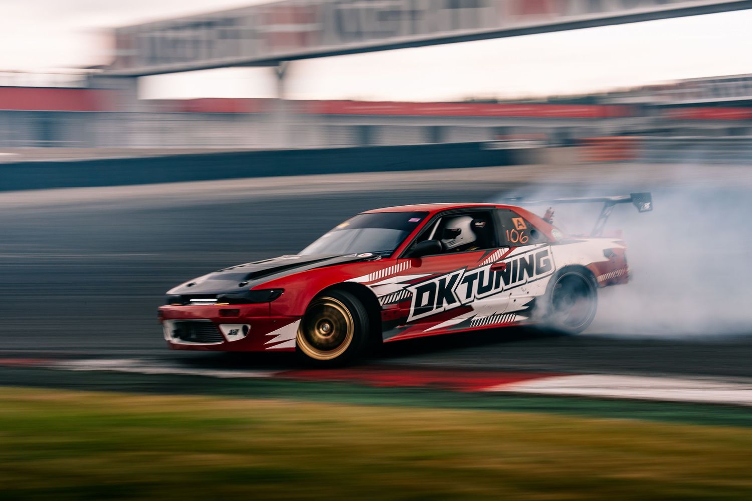 A red sports car drifts sideways on a racetrack creating smoke from burning rubber during a drift competition.