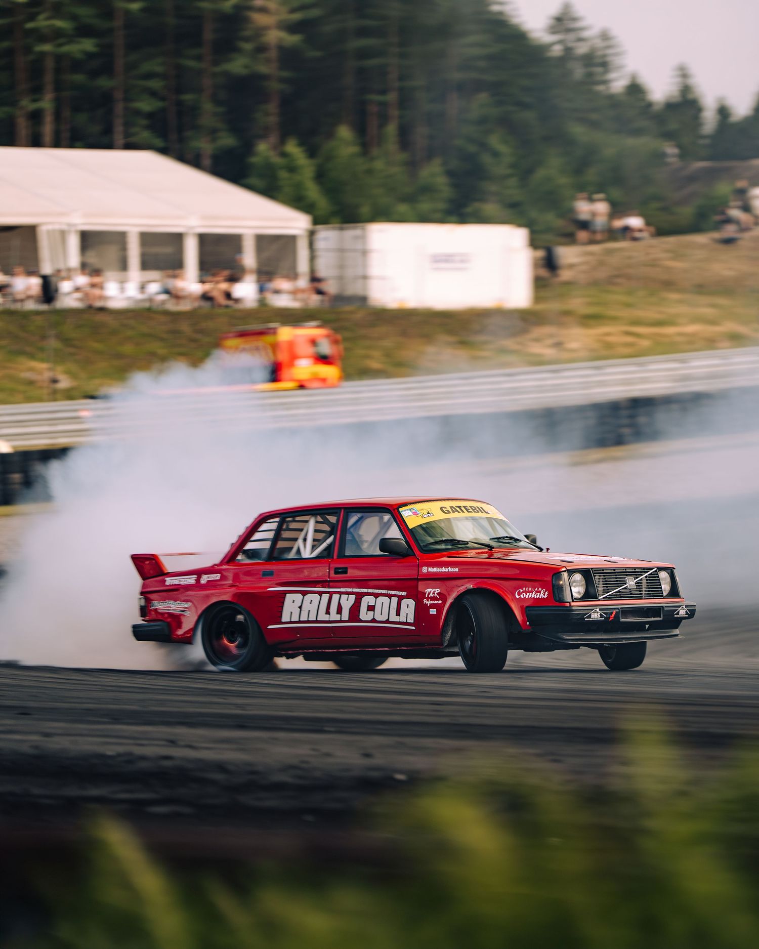 A red vintage Volvo rally car drifts sideways creating smoke on a racetrack with buildings and forest in the background.
