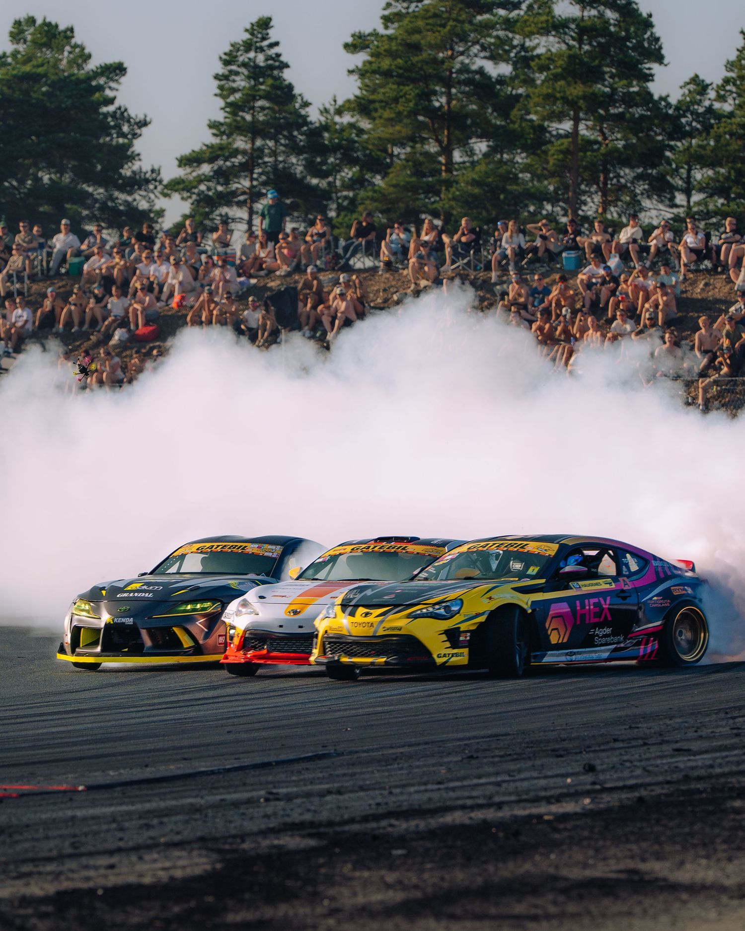 Multiple colorful drift cars perform synchronized drifting on a track with smoke trails and a large crowd watching.