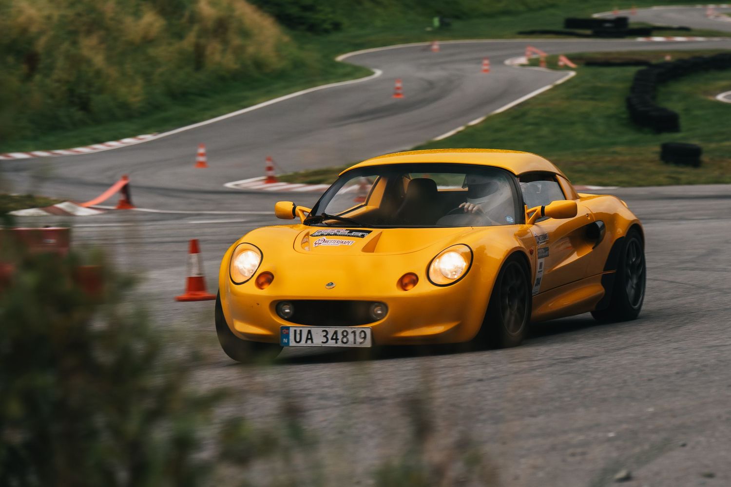 A yellow Lotus Exige sports car races around a winding track with orange cones marking the course.