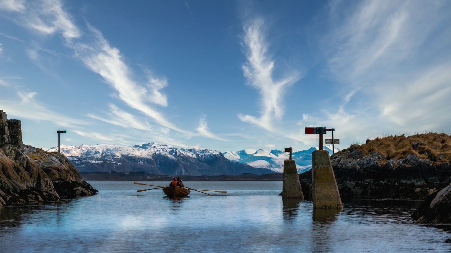 Navigation markers stand in tranquil fjord waters with snow-capped mountains in the distance.