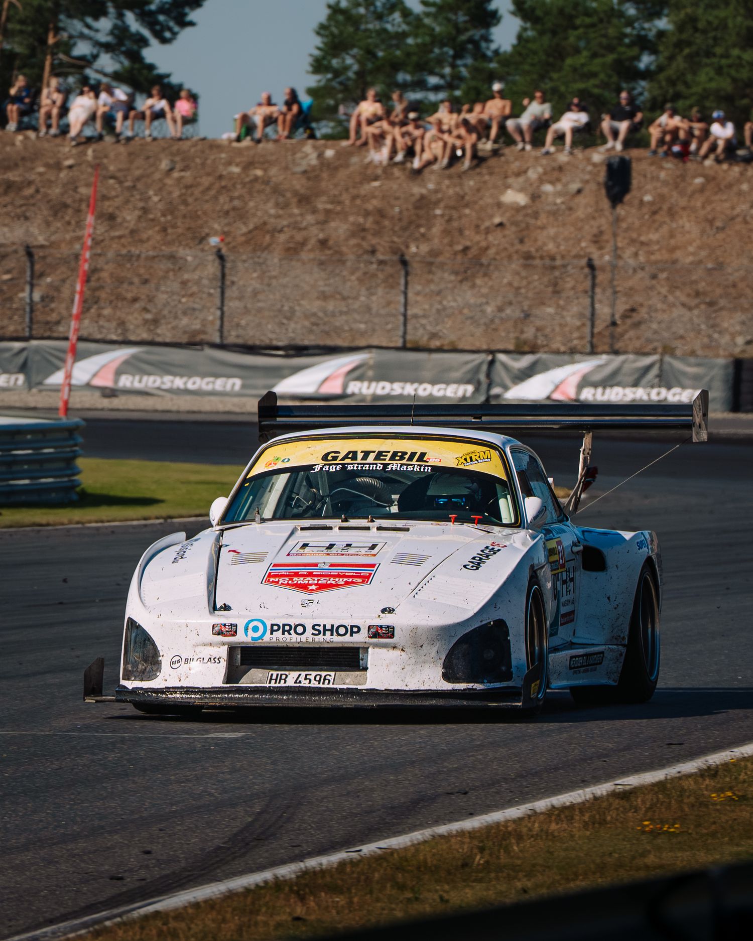 A white racing car navigates a turn on a racetrack with spectators watching from a hill in the background.
