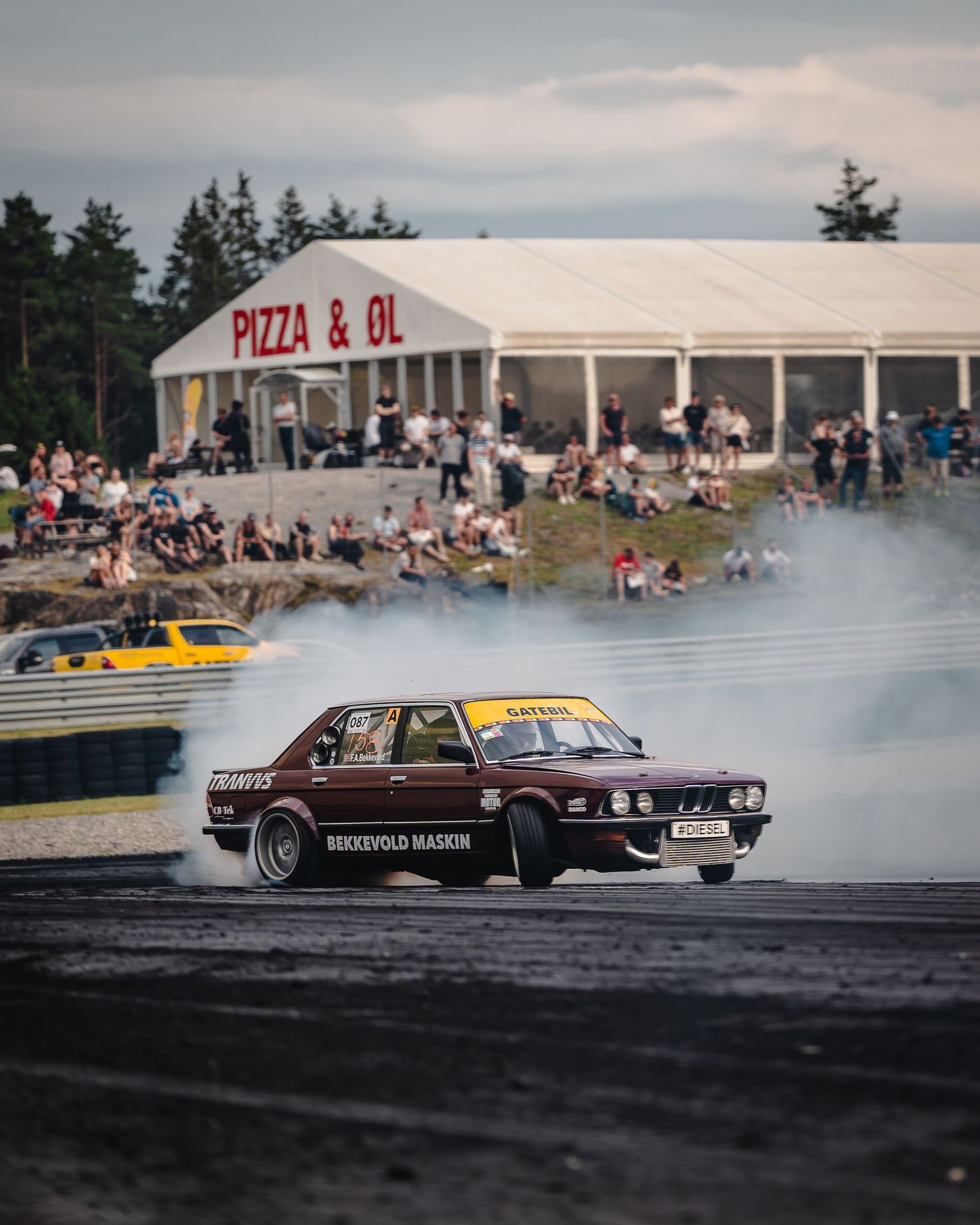 A brown vintage car drifts sideways on a race track creating smoke during a drift competition event.