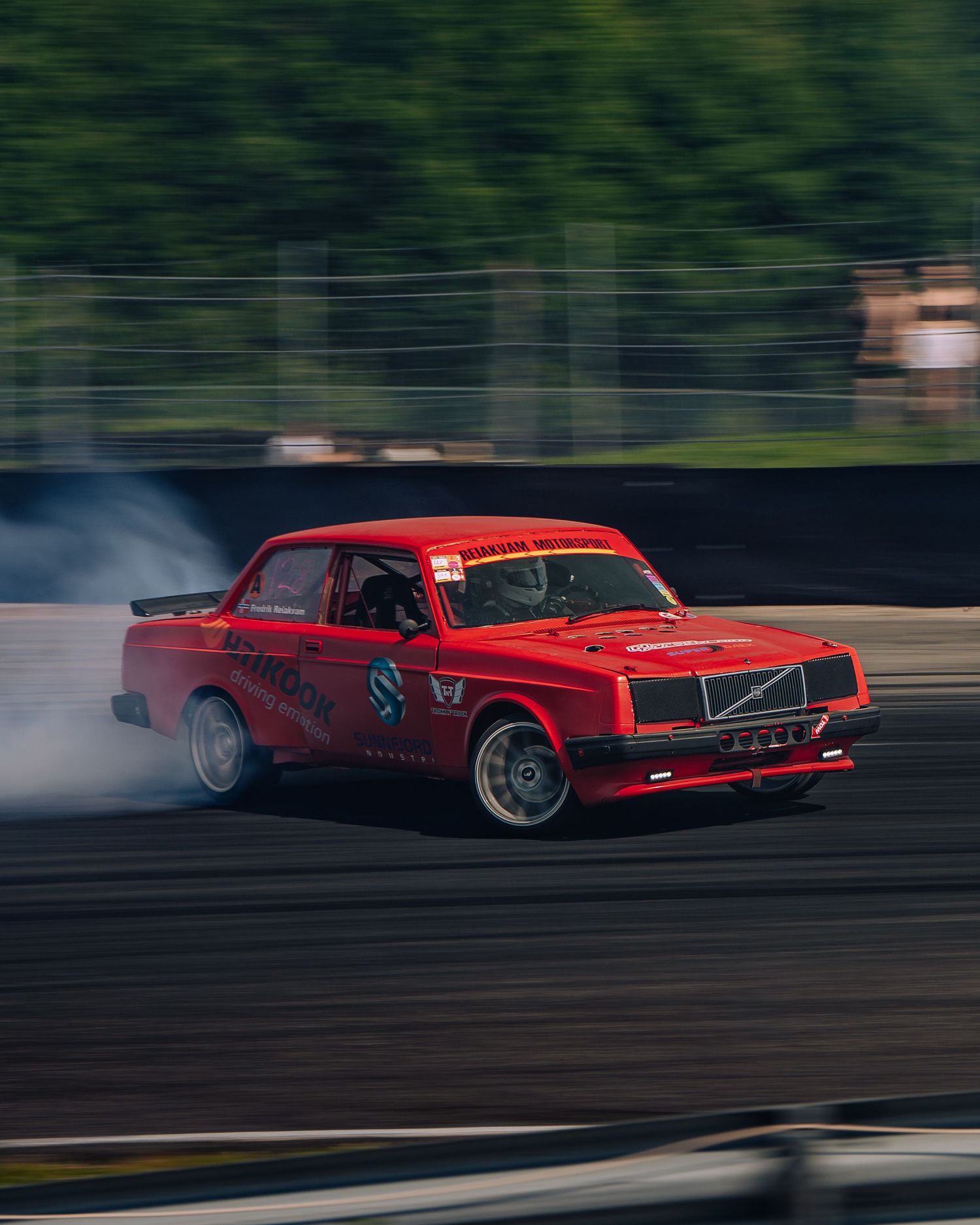 Red Volvo drift car sliding sideways on a race track creating tire smoke during a drifting competition.