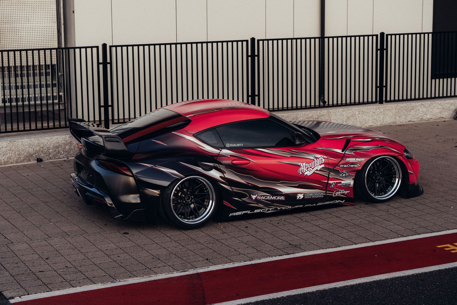 A modified red and black sports car with wide body kit and custom graphics parked on a street near metal fencing.