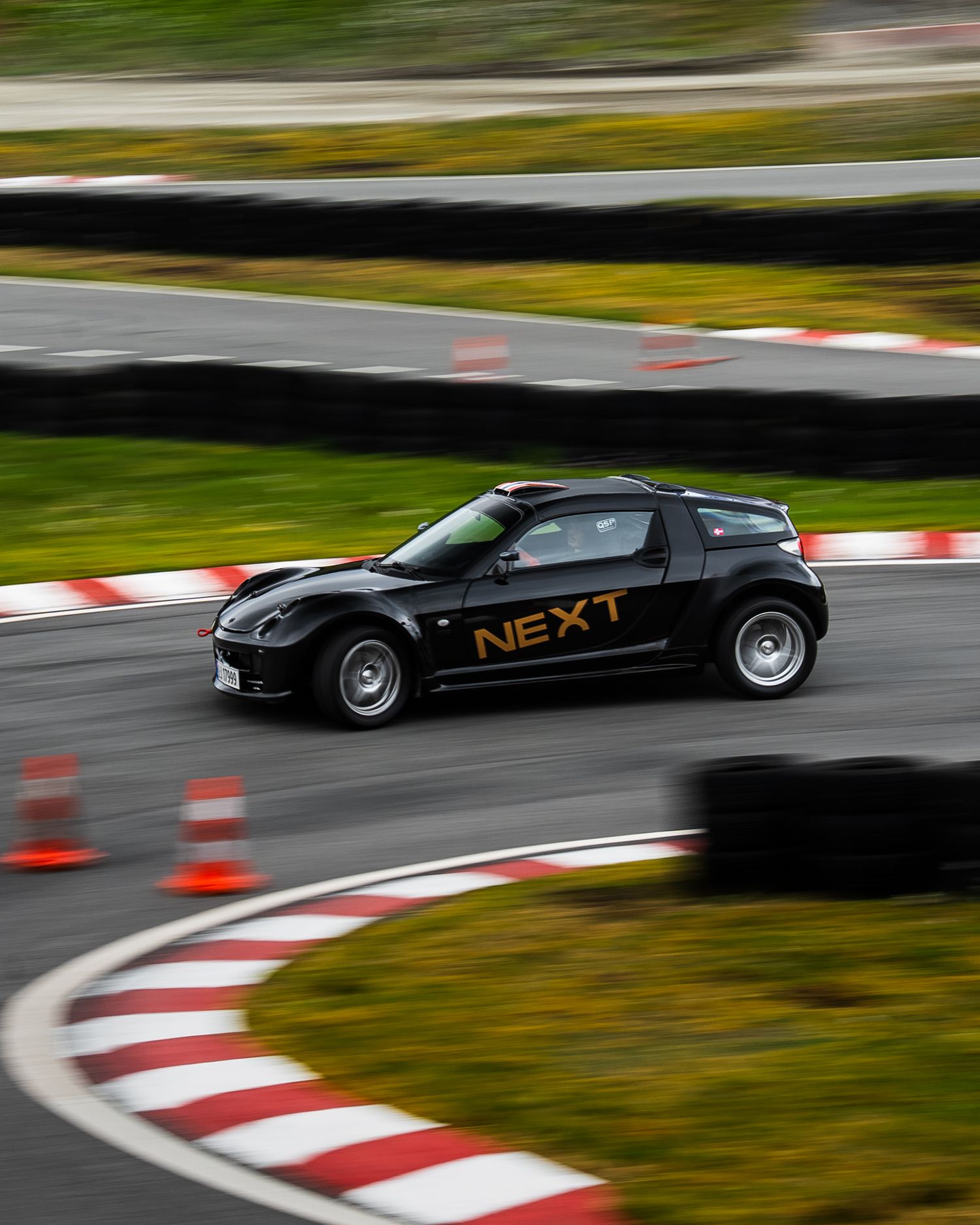 Black sports car with NEXT branding racing around a curved track with red and white curbing and safety cones.