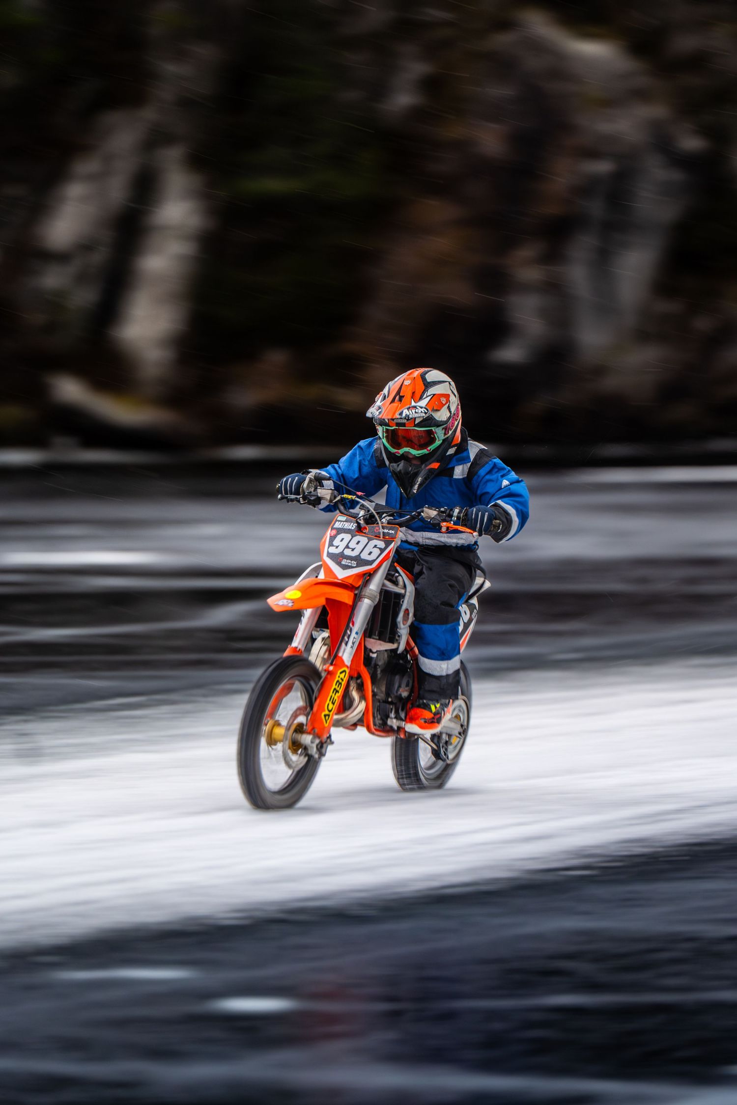 Motorcycle rider races across a frozen lake surface with snow spray trailing behind the orange dirt bike.