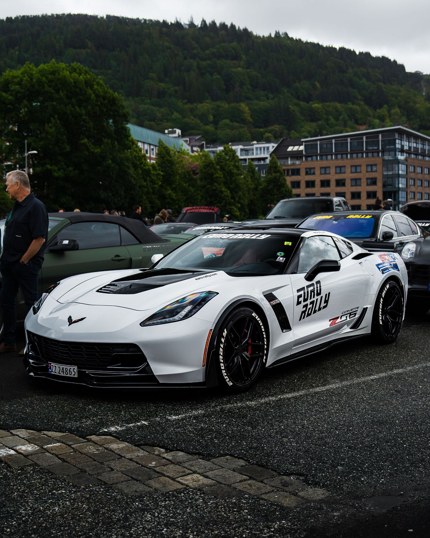 White Chevrolet Corvette racing car with custom decals parked in a wet parking lot with hills in the background.
