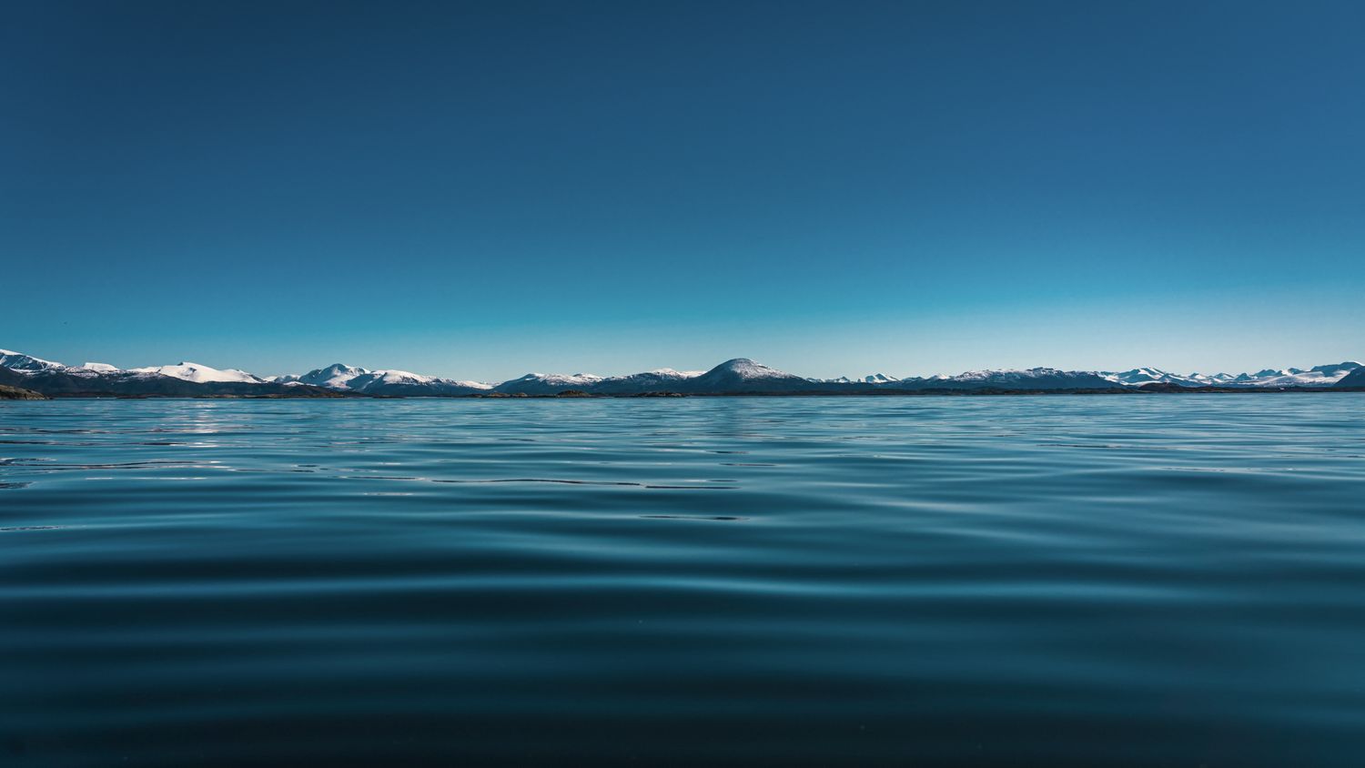 Calm blue waters stretching to snow-capped mountain range under clear sky.