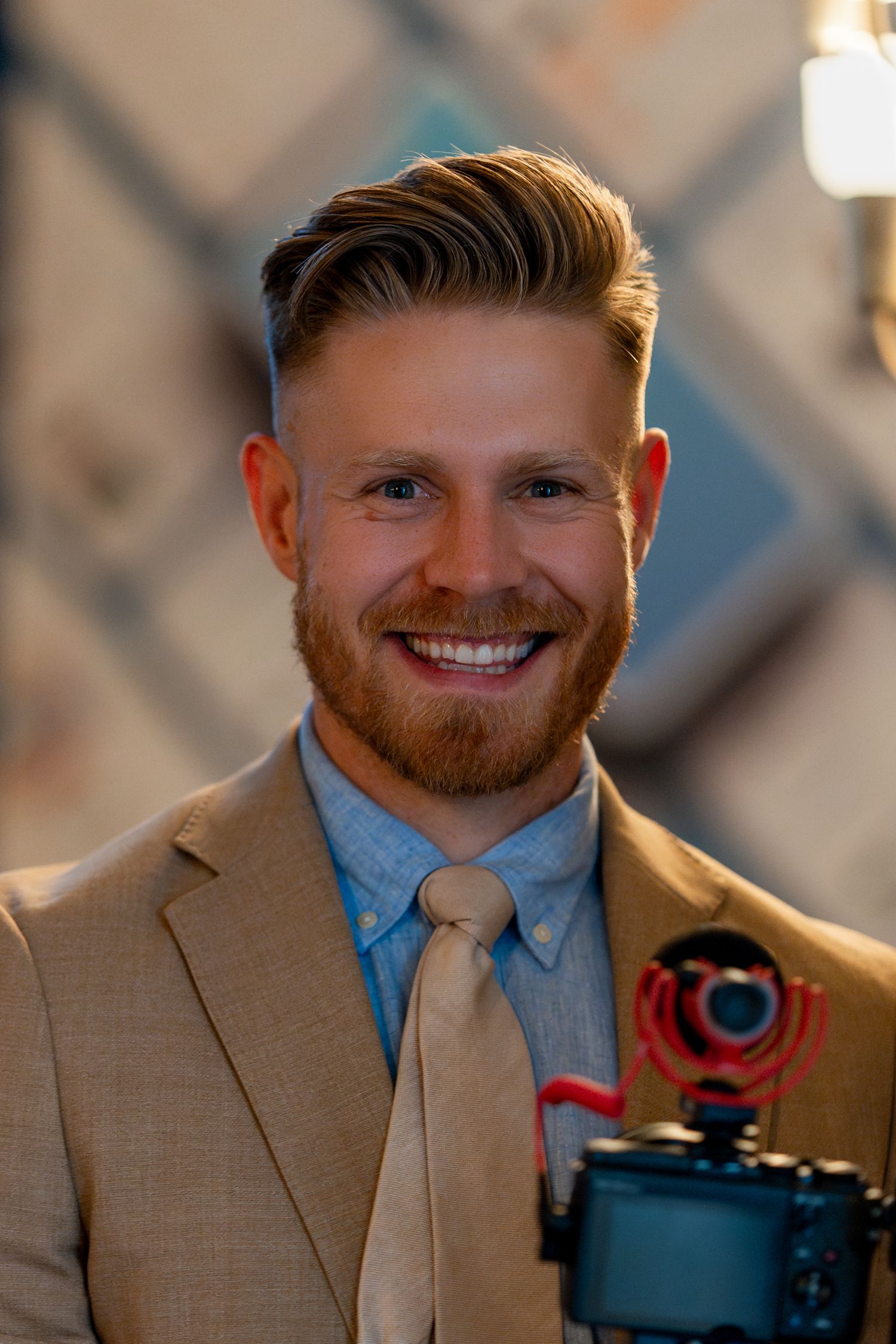 Professional with styled hair and beard smiles while holding camera equipment.