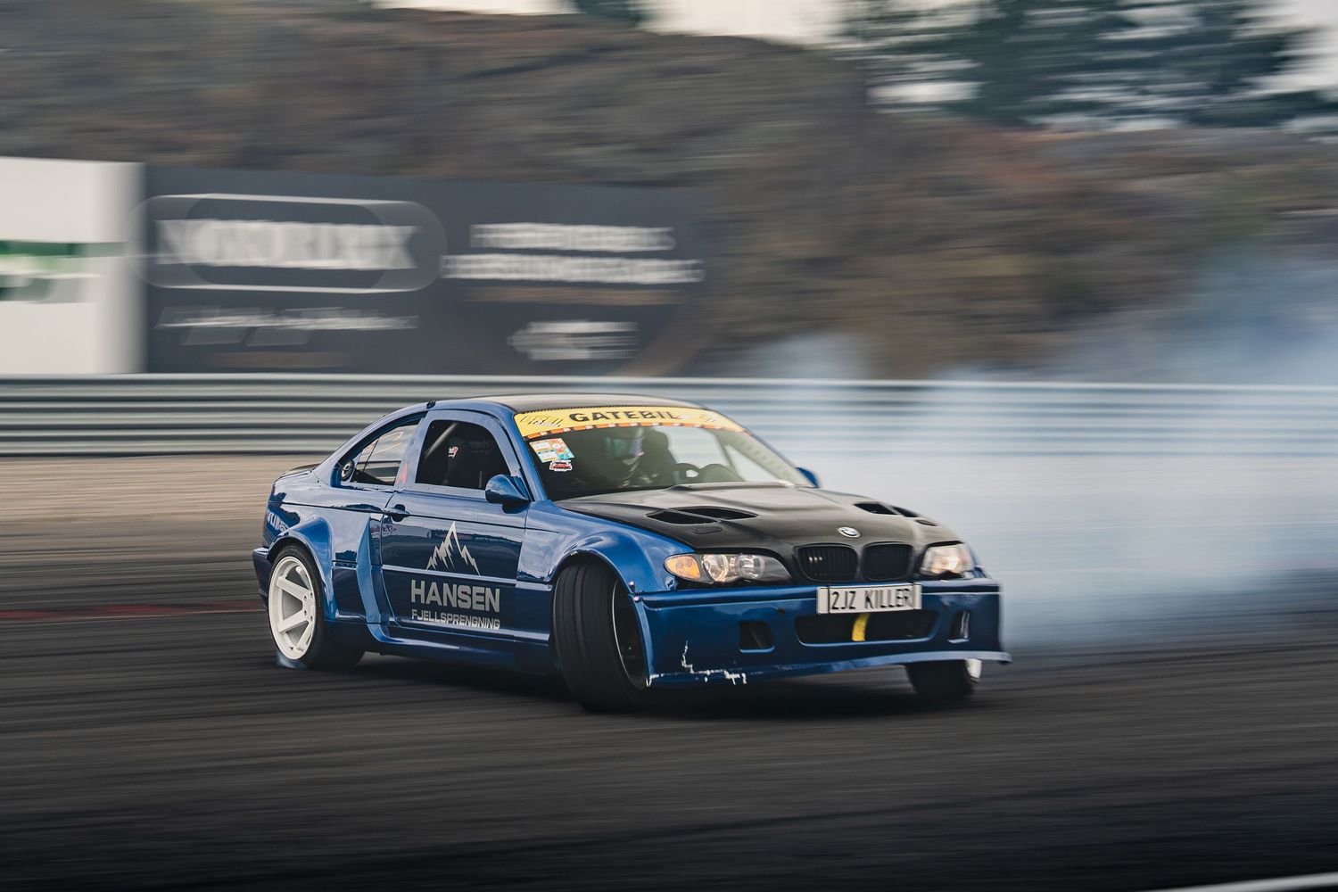 A series of drifting cars creating smoke clouds during a motorsport competition on a racetrack.