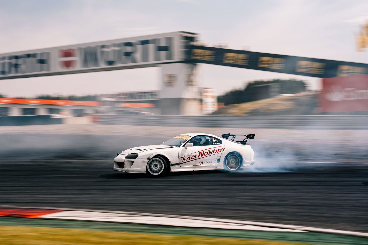 Multiple racing cars drift around a track with smoke trailing behind them during a motorsport competition.