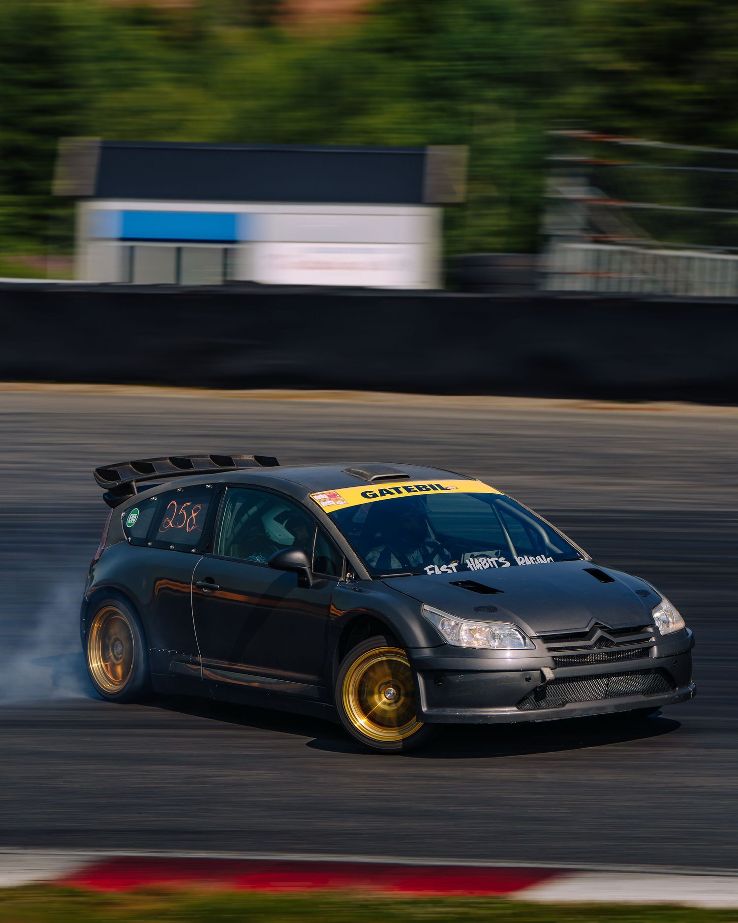 A group of modified sports cars drifting in tandem on a race track with smoke trailing behind their tires.