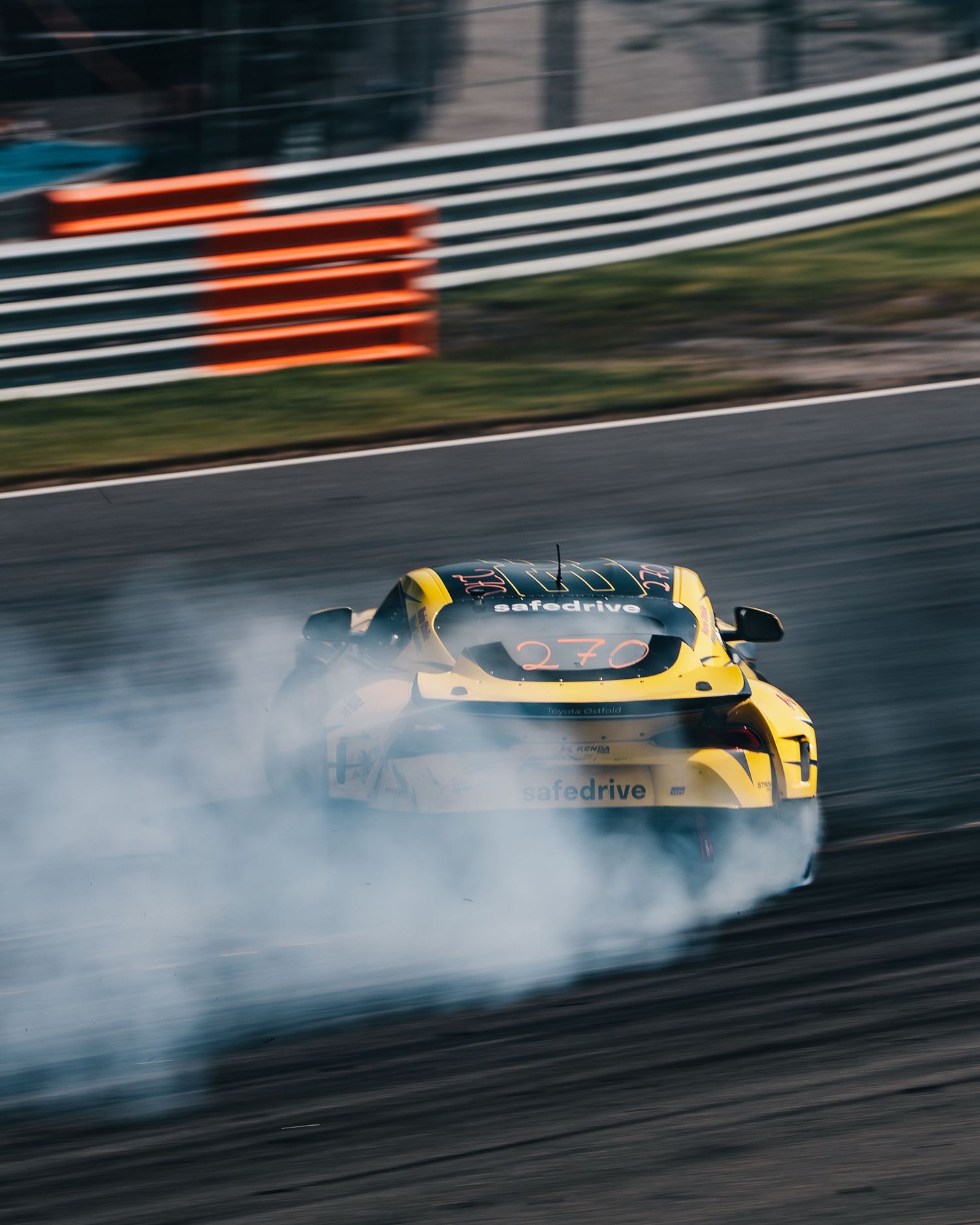 Yellow drift car sliding sideways on racetrack with smoke trailing from spinning tires during competition.