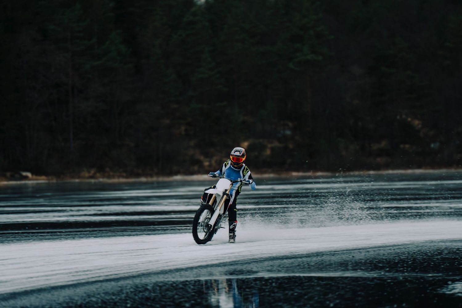A motorcycle rider performs a stunt riding across a frozen lake surface in a dark forest setting.