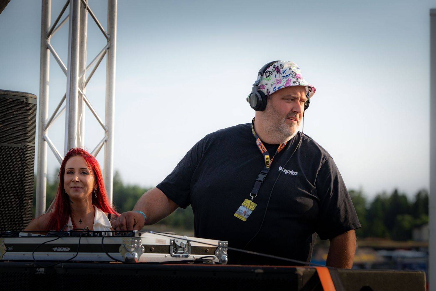DJ wearing bandana and black t-shirt performs on stage at outdoor event while woman with red hair looks on in background.