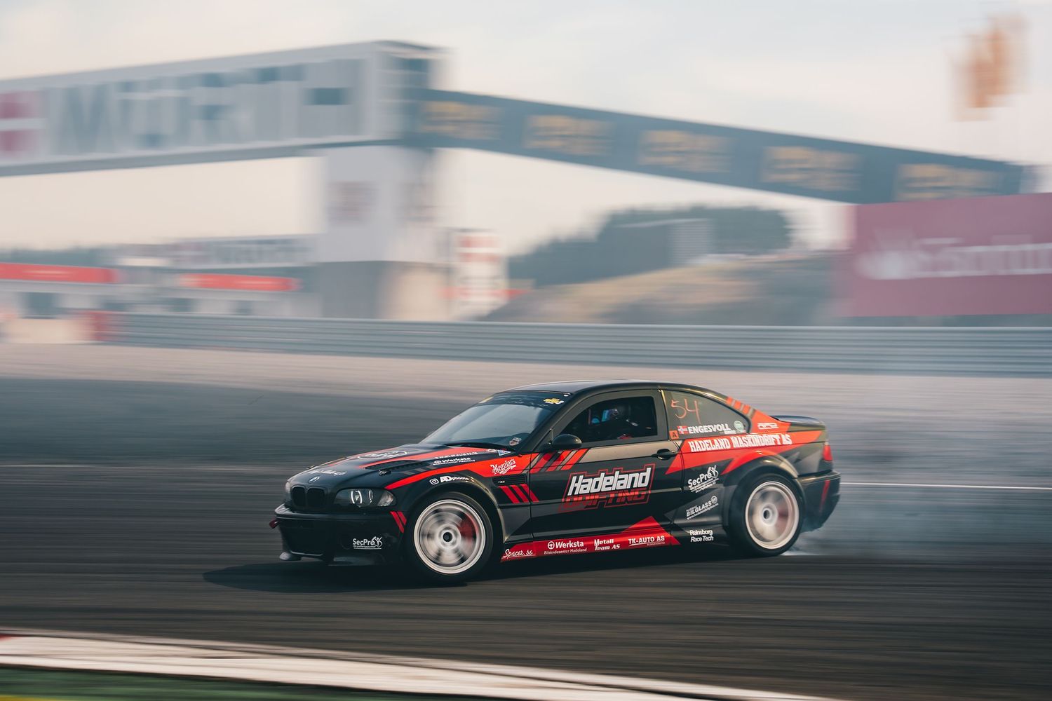 A red and black drift car slides sideways around a racetrack with smoke billowing from its tires during competition.