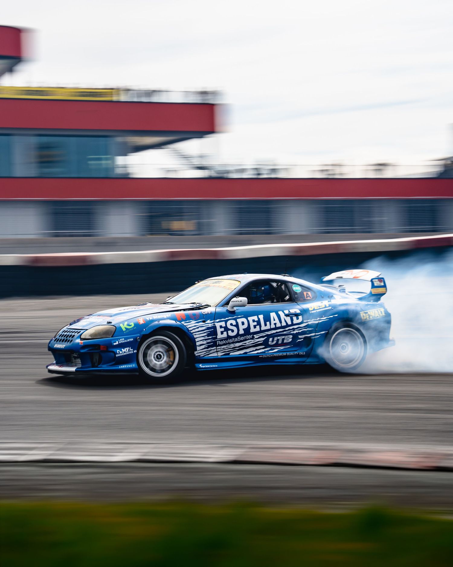 A blue drift car slides sideways creating smoke during a motorsport competition on a racetrack.