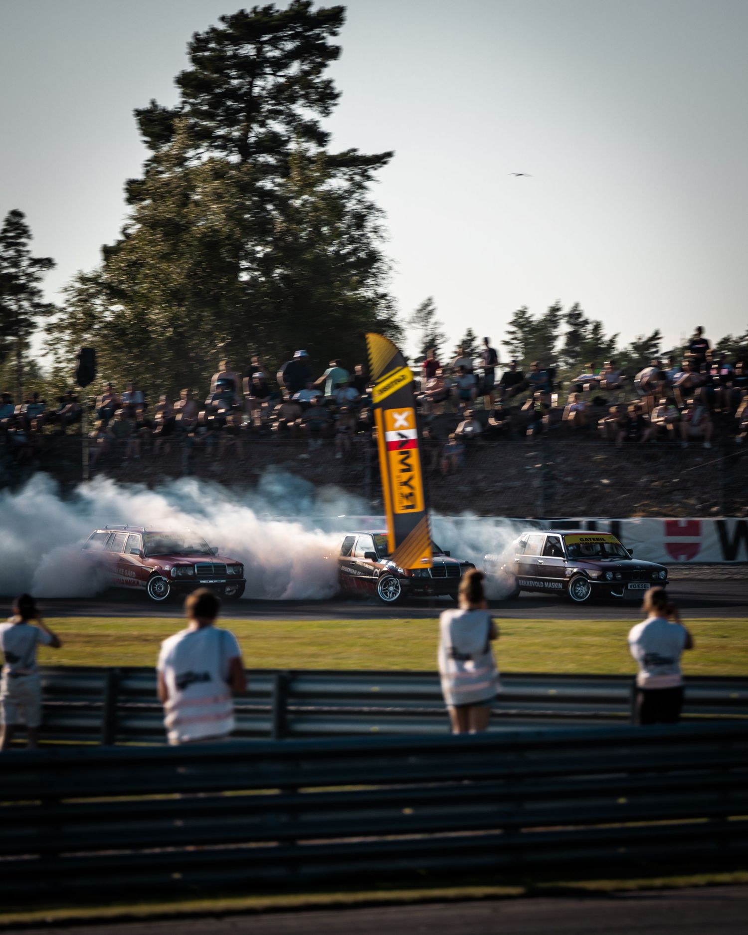 Drift cars perform synchronized burnouts on a racetrack as spectators watch from behind barriers at sunset.