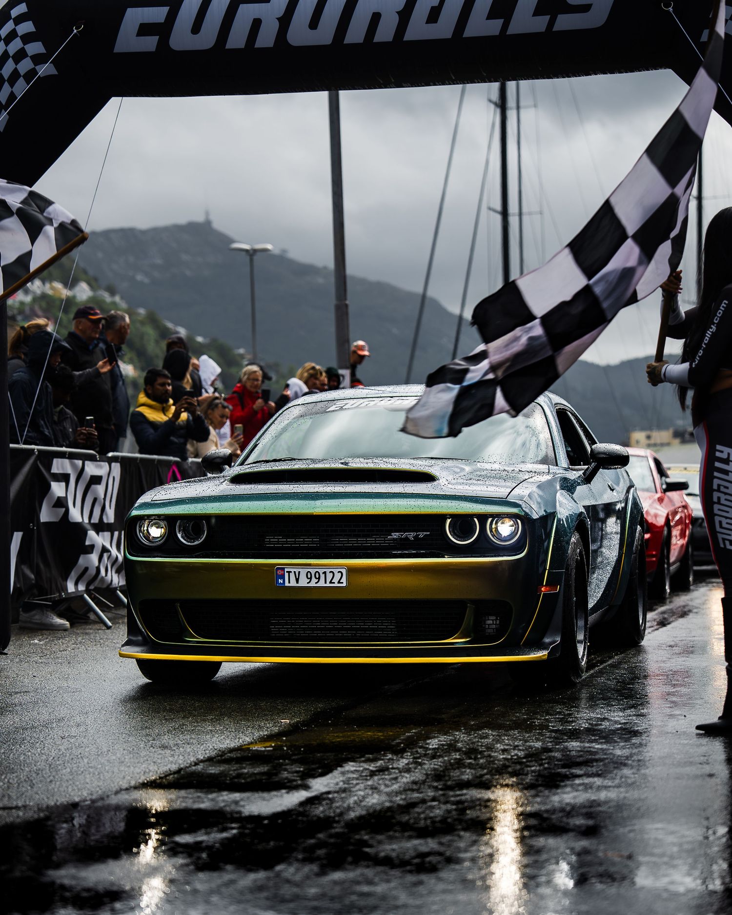 Dark green Dodge Challenger Hellcat crosses Eurorally finish line on rainy mountain track.