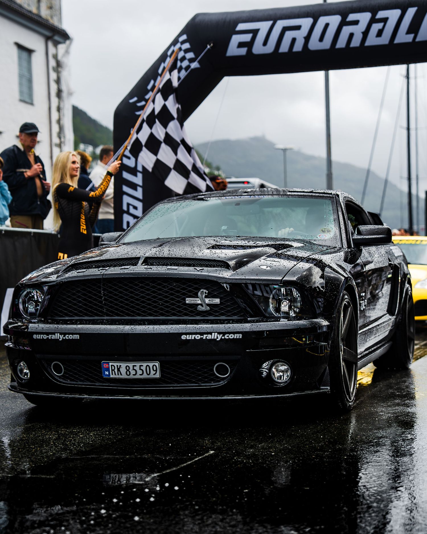 Black Shelby Mustang crosses finish line under Eurorally arch on wet racing track with mountains in background.