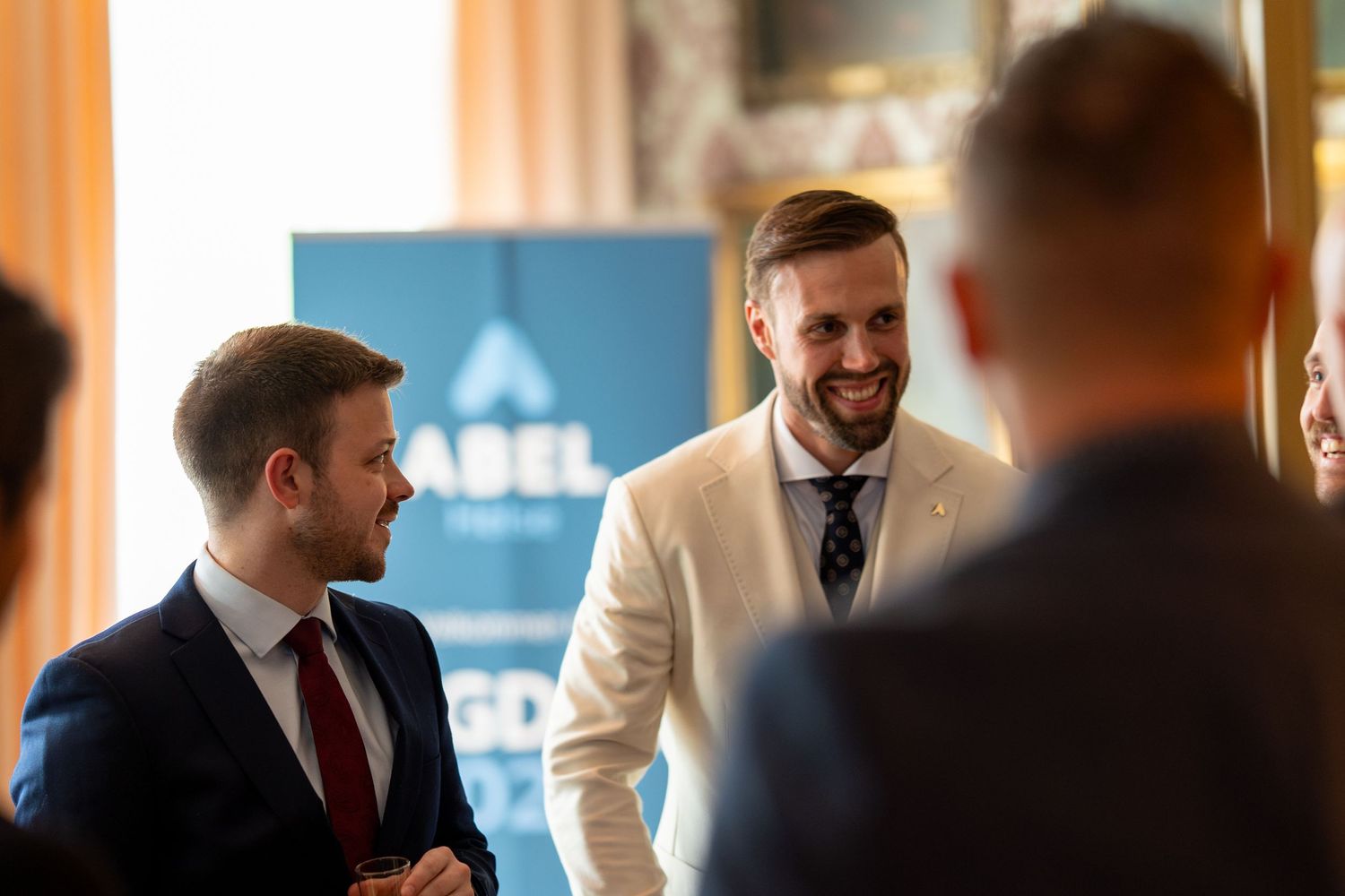 Business professionals engage in animated conversation at a corporate event with blue branding visible in background.