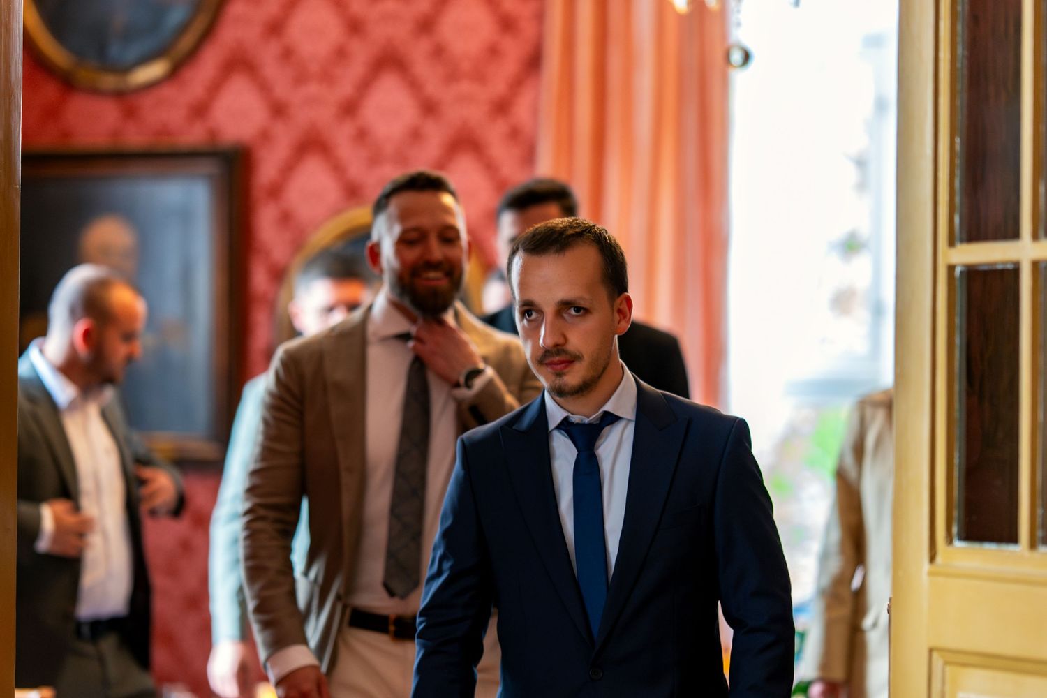 Guests mingling in formal attire at elegant venue with ornate red wallpaper.
