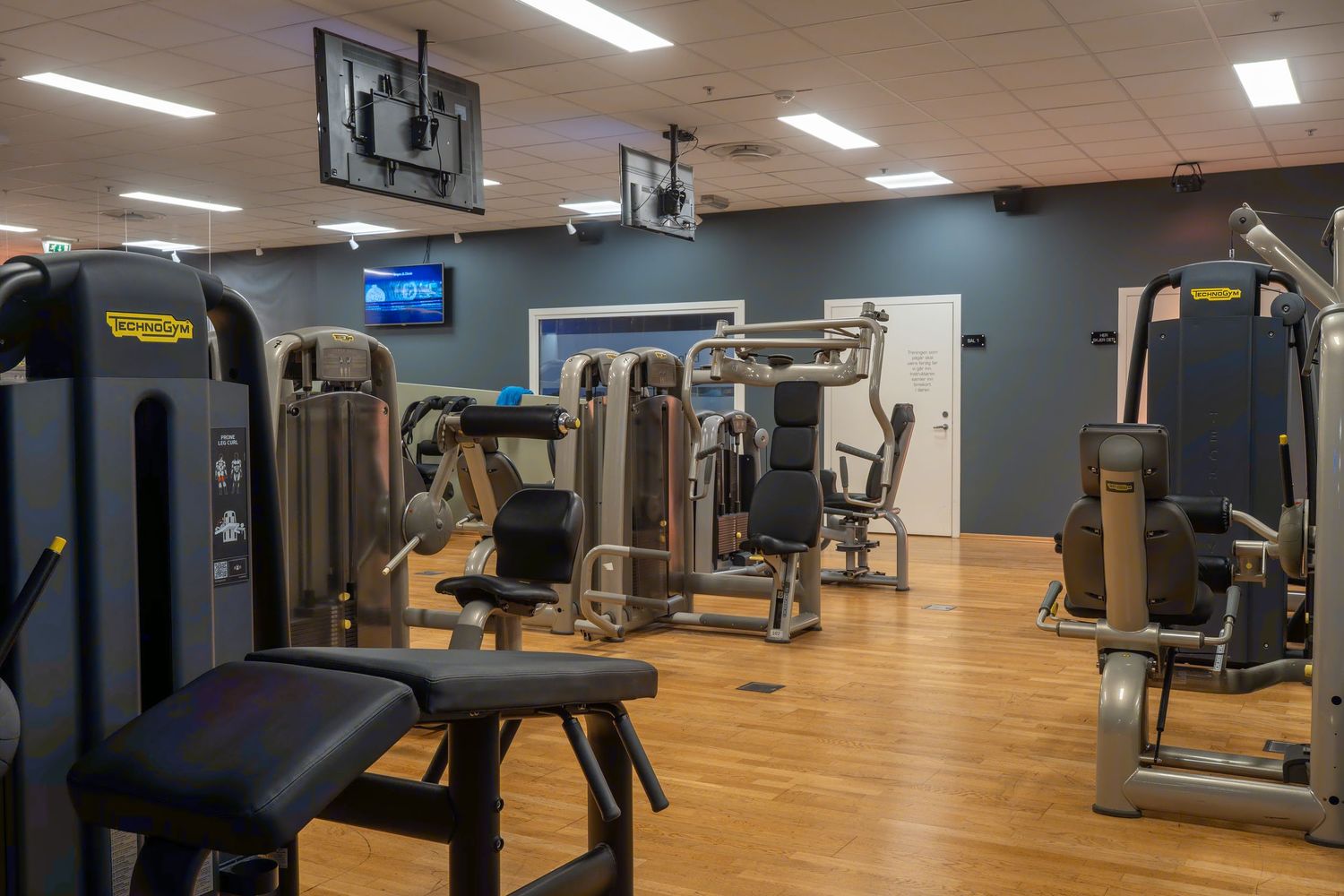 Modern fitness center with treadmills and exercise equipment arranged in rows on hardwood flooring under bright lighting.