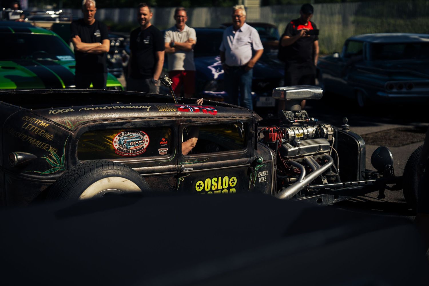 Custom hot rod with exposed engine displayed at dusk during car meet.