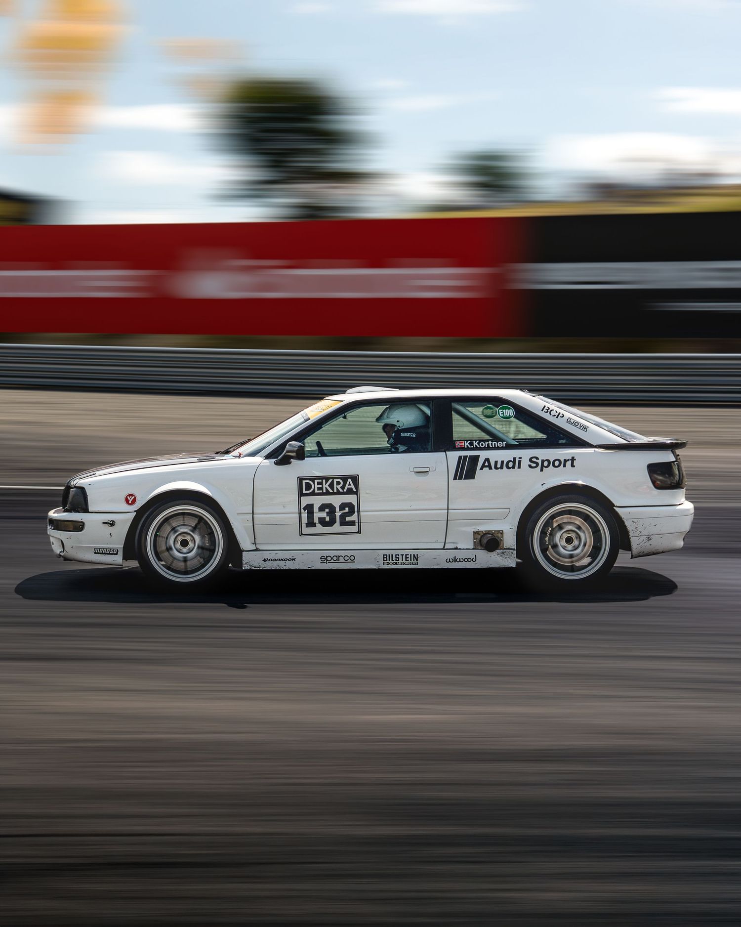 White race car with Audi Sport branding speeds along a racetrack creating a motion blur effect.