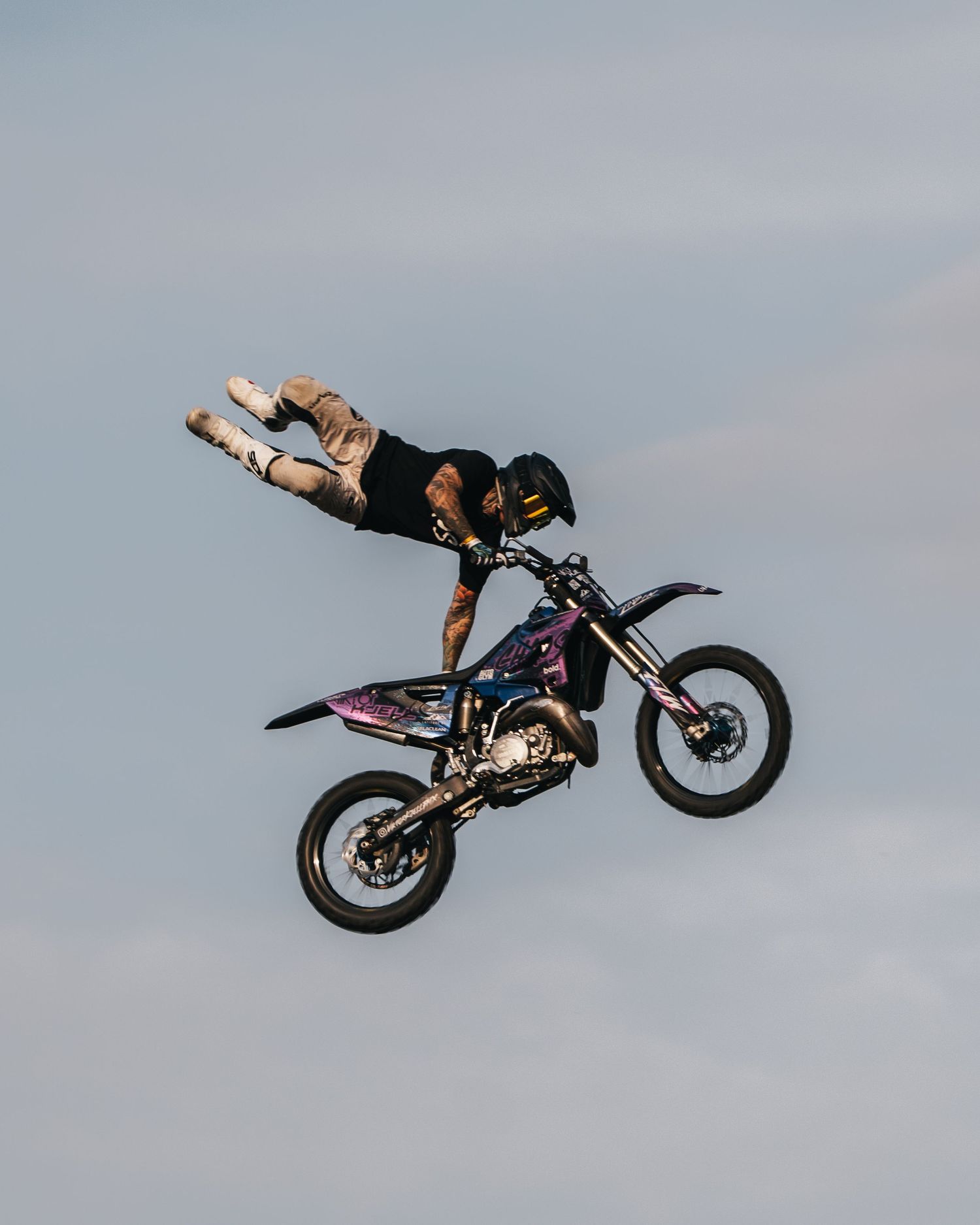 Motorcycle rider performs a dramatic backflip stunt against a cloudy sky backdrop during an extreme sports event.