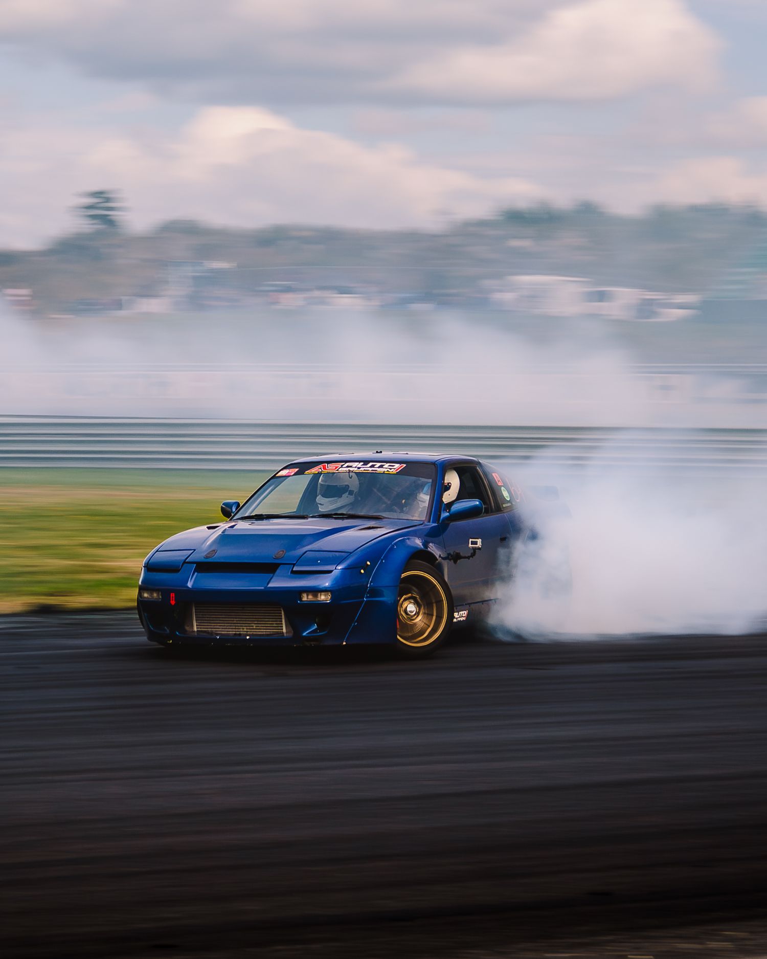 A blue sports car performs a dramatic drift with smoke billowing from its tires on a race track.