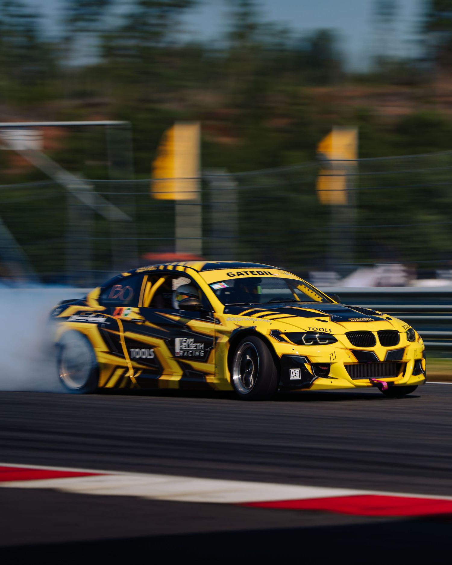 Yellow BMW race car drifts sideways on track creating smoke during competitive motorsport event.